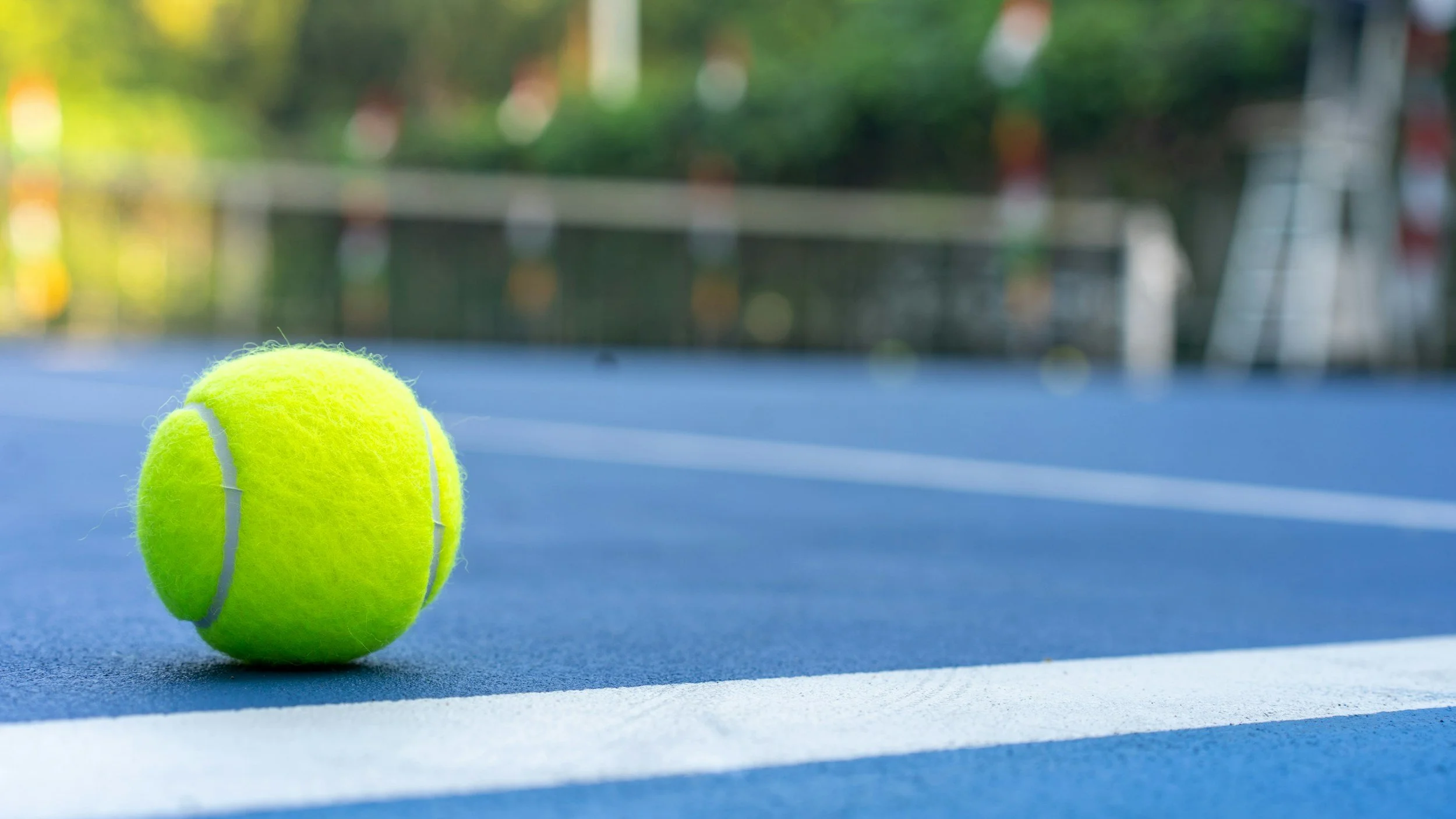 A bright yellow tennis ball on a blue tennis court with white lines. The background includes blurred greenery and outdoor sports equipment.