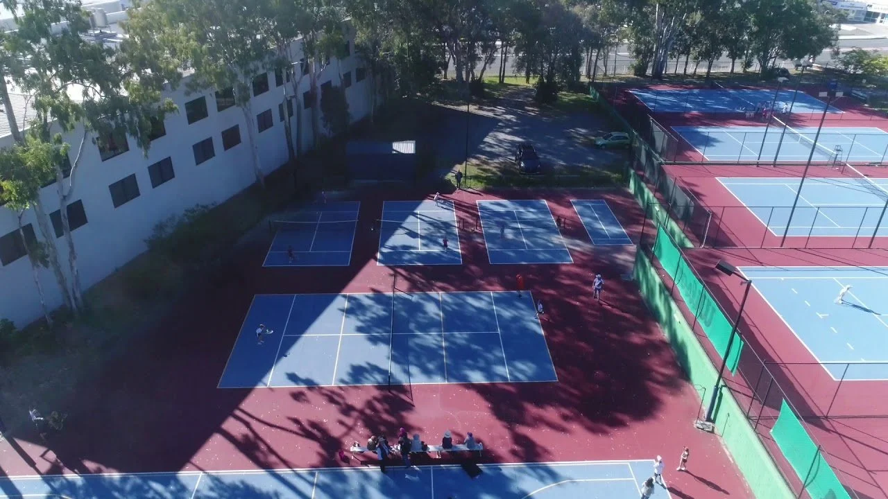 Aerial view of a tennis and pickleball court complex with players on the courts and trees casting shadows.