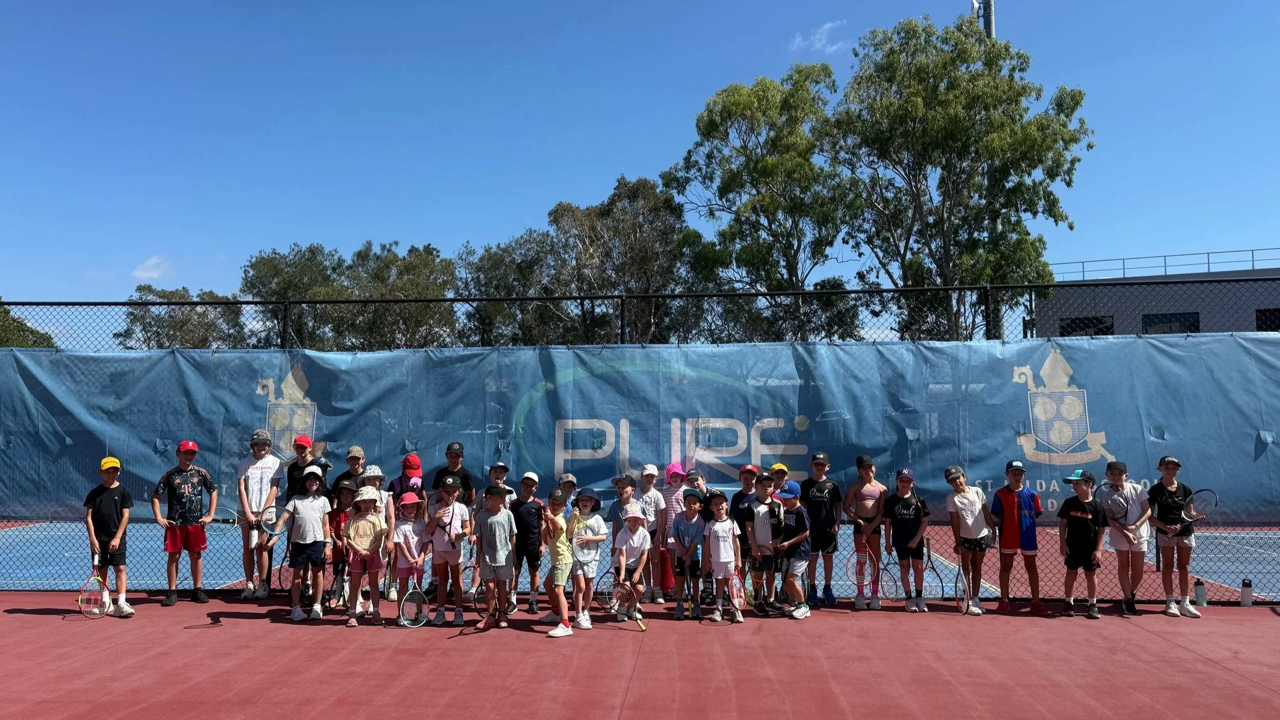 Group of children and coaching staff standing on a tennis court, holding tennis rackets, with a blue banner and trees in the background on a sunny day.
