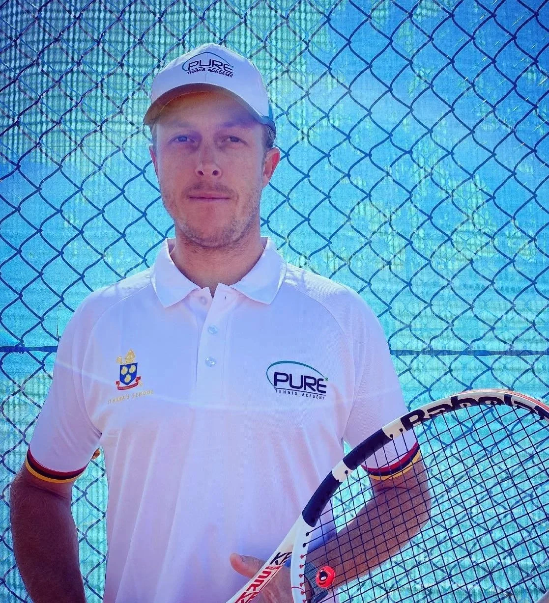 A man in a white polo shirt and cap standing in front of a tennis court, holding a tennis racket. The shirt and cap have the 'PURE TENNIS ACADEMY' logo. There is a blue chain-link fence behind him.