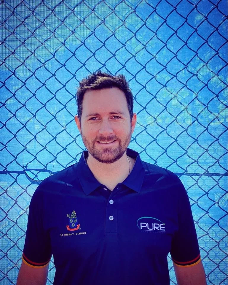 A young man with short brown hair and a beard smiling, standing in front of a chain-link fence.