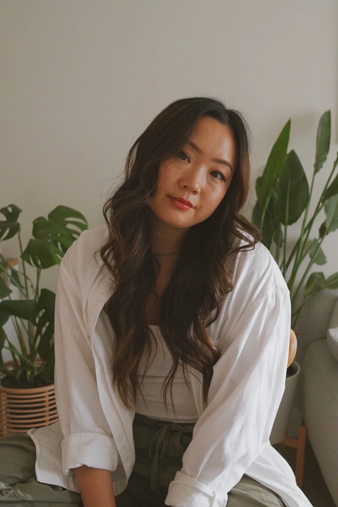 A woman with long wavy dark hair, wearing a white shirt and olive green pants, sitting indoors with houseplants in the background.
