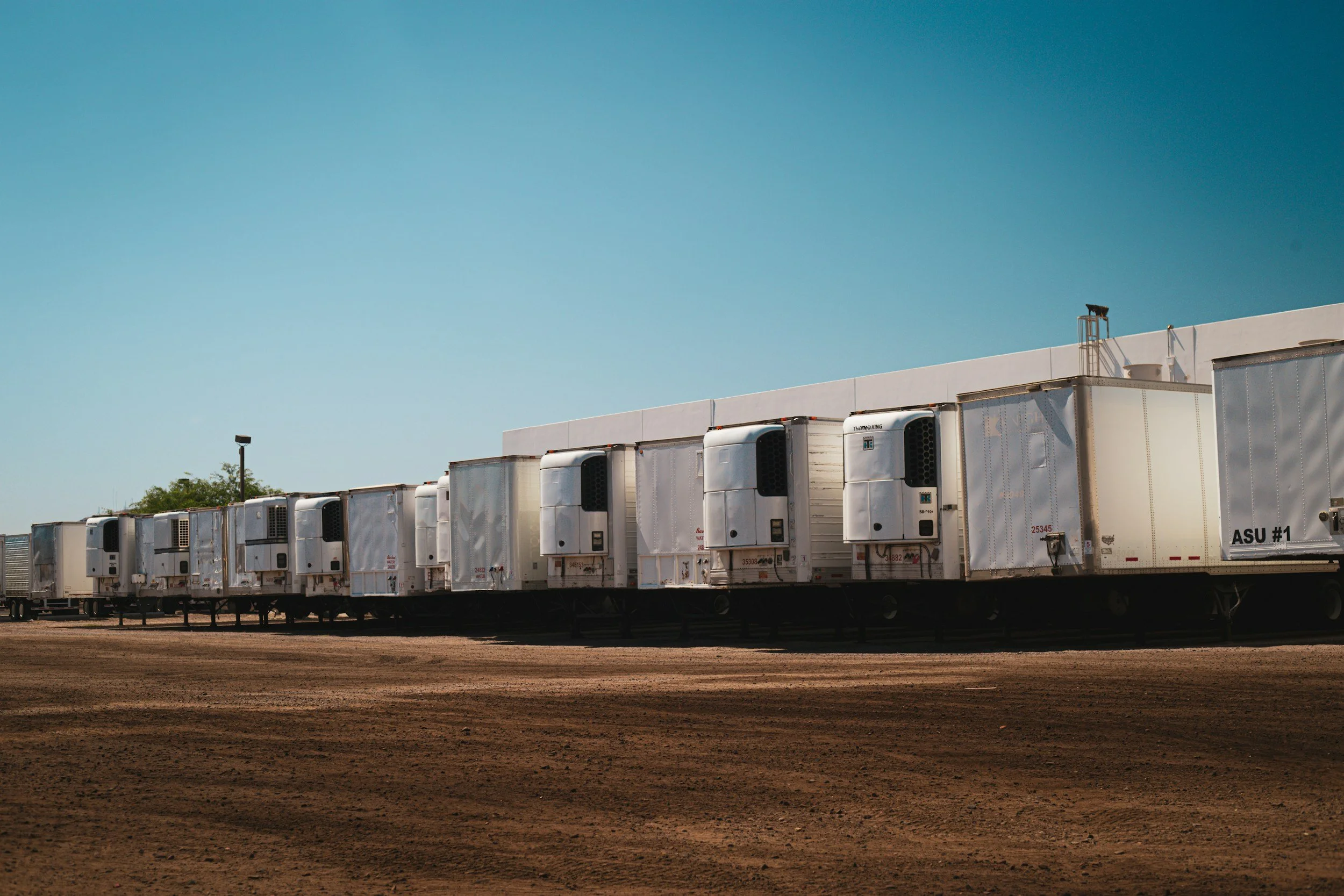 A row of white semi-trailers parked on a dirt lot under a clear blue sky.
