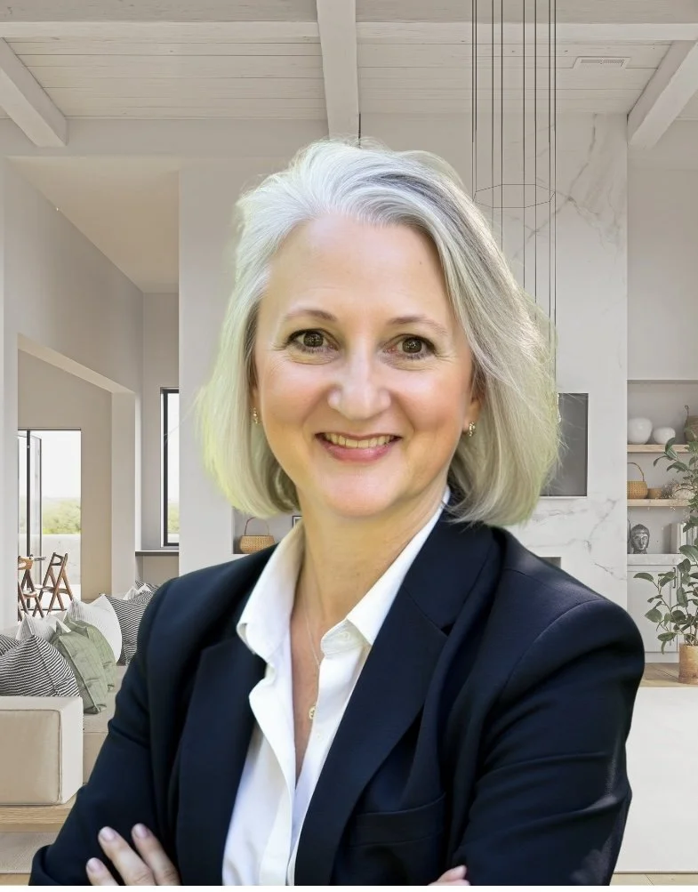 A woman with shoulder-length gray hair smiling with her arms crossed, wearing a navy blazer and a white shirt, standing in a bright, modern living room with large windows, white walls, and minimalistic decor.
