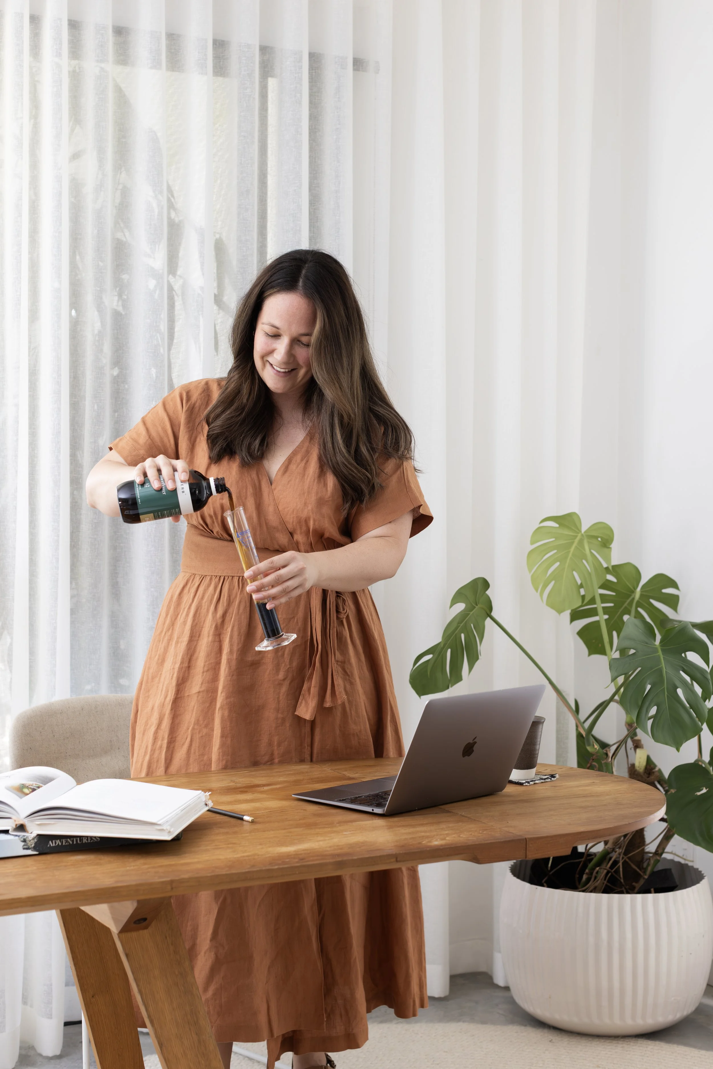 A woman with long brown hair, wearing a brown dress, is smiling while pouring a dark beverage into a champagne flute in a bright room with white curtains. There is a laptop, open books, and a potted plant on a wooden table.