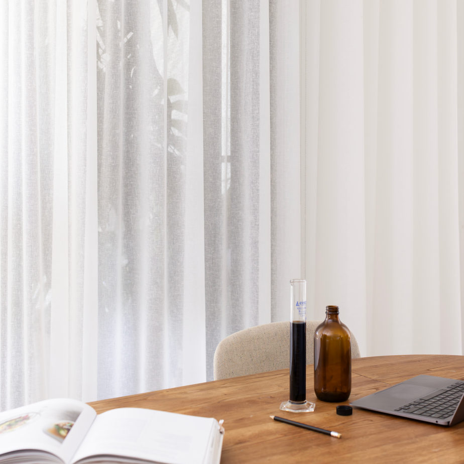 A wooden table with an open book, a laptop, a black pen, a small black cap, a tall graduated cylinder with a dark liquid, and an amber glass bottle, set against sheer white curtains.