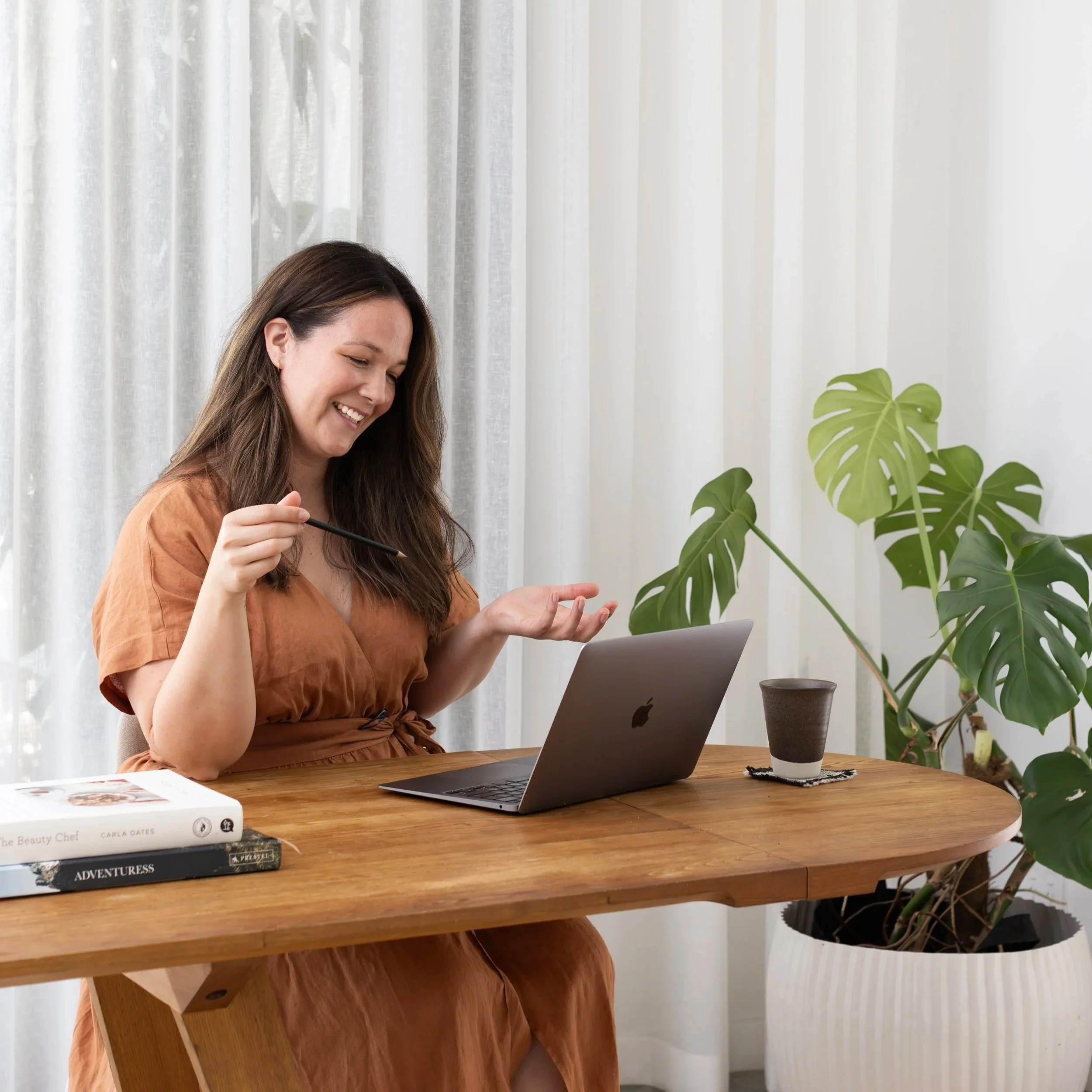 A woman sitting at a wooden table, smiling at her laptop, with house plants and books in the background.