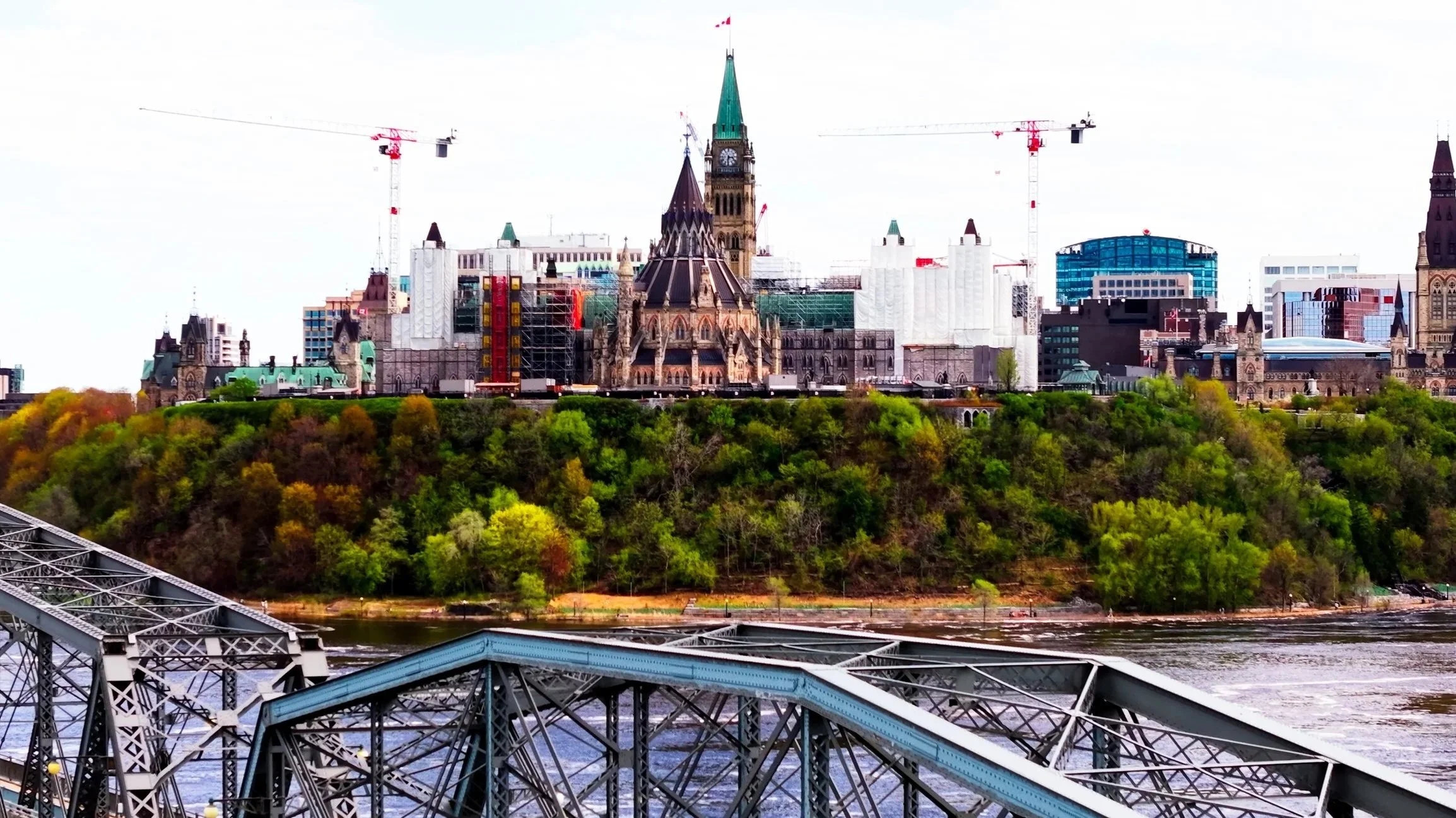 View of Parliament Hill in Ottawa, Canada, with historic buildings, green trees, and a river in the foreground.