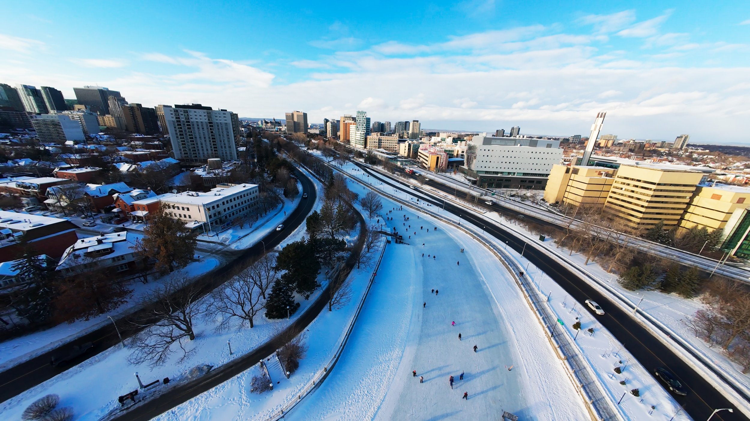 Aerial view of a snowy urban park with people ice skating, surrounded by city buildings and roads on a clear winter day.