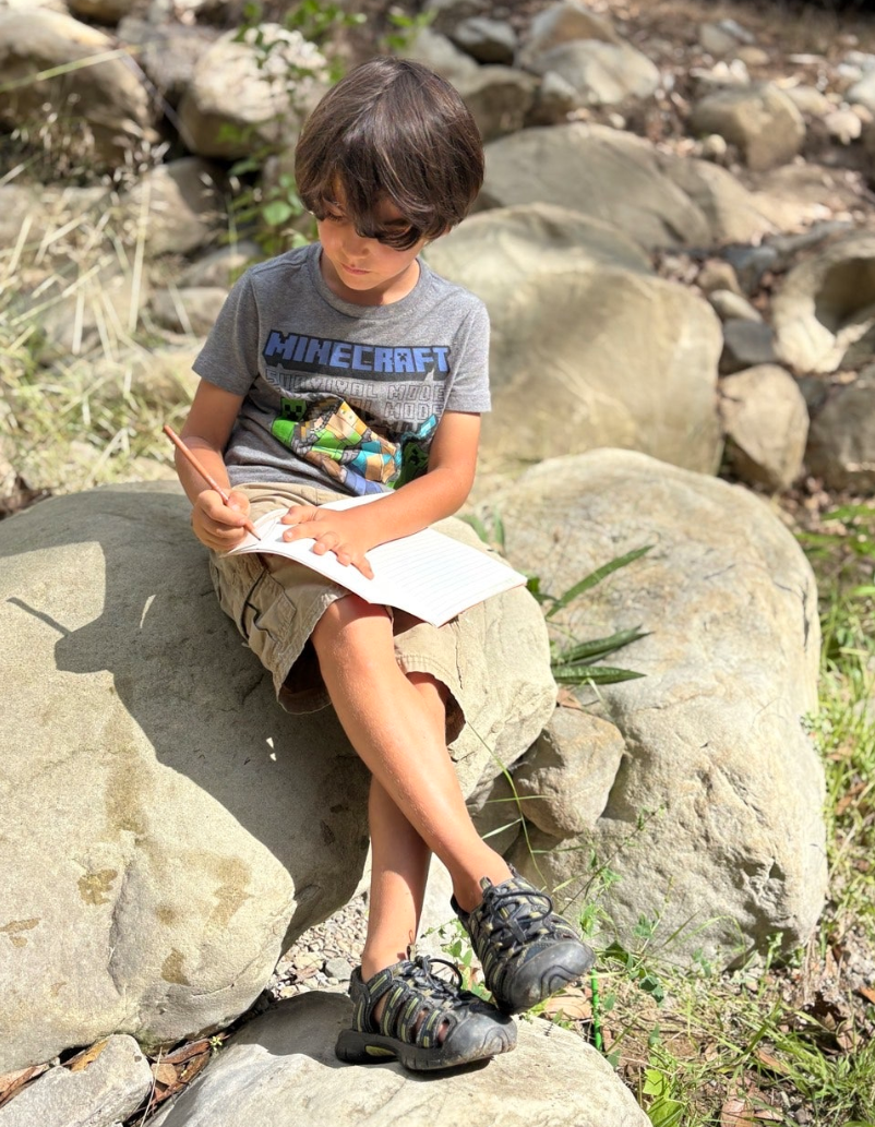 A young boy sitting on a large rock outdoors, writing in a notebook with a pencil. He is wearing a gray Minecraft T-shirt, tan shorts, and hiking shoes. There are several rocks and some green plants around him.