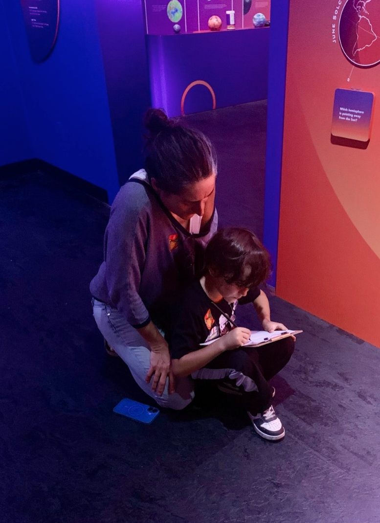 A woman and a young boy sitting on the ground in a science museum, looking at a notebook. The scene is illuminated with purple and orange lighting.