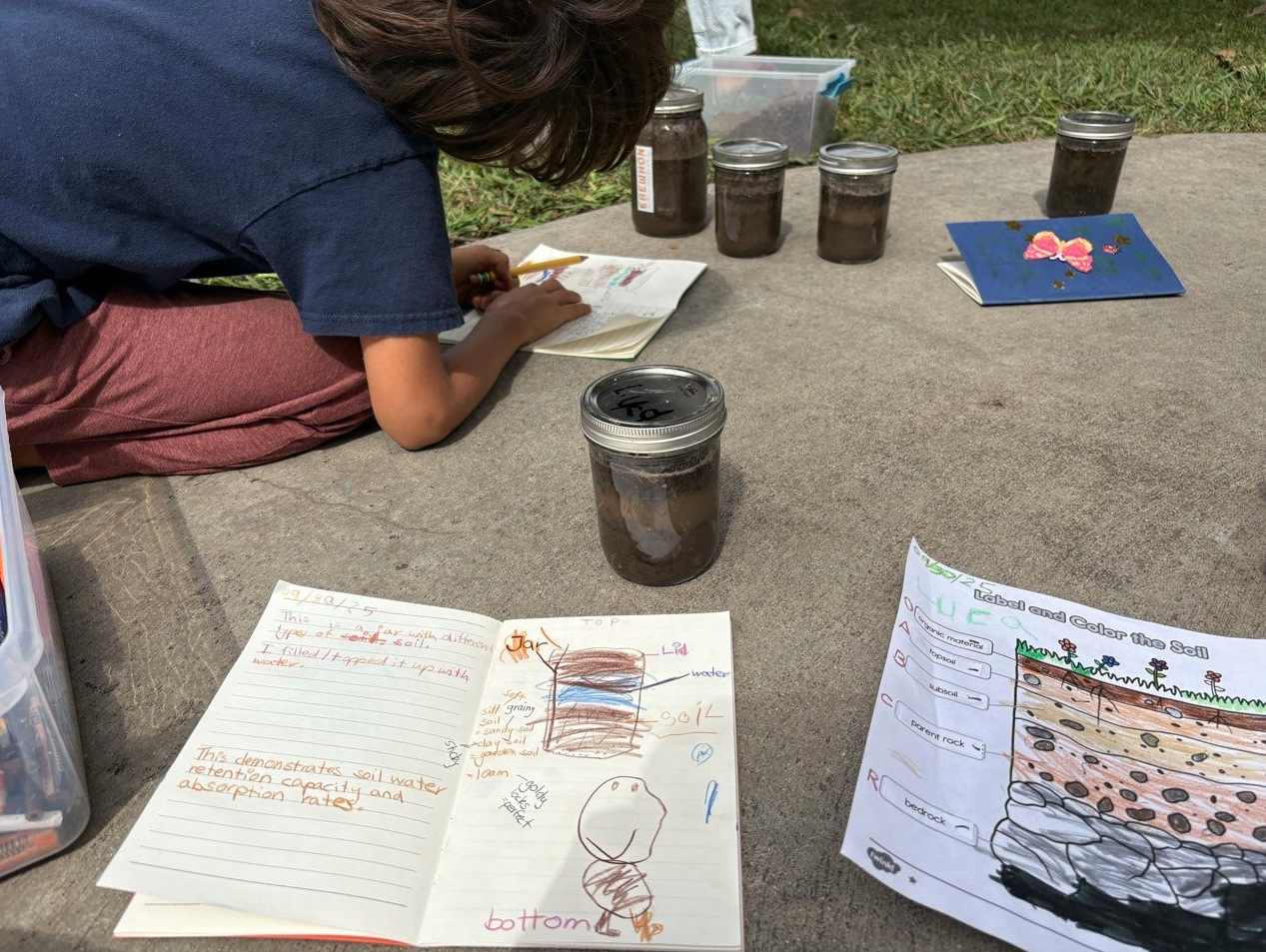 Child kneeling on a concrete surface, writing in a notebook with jars of soil and a diagram about soil layers nearby, outdoors on a grassy area.