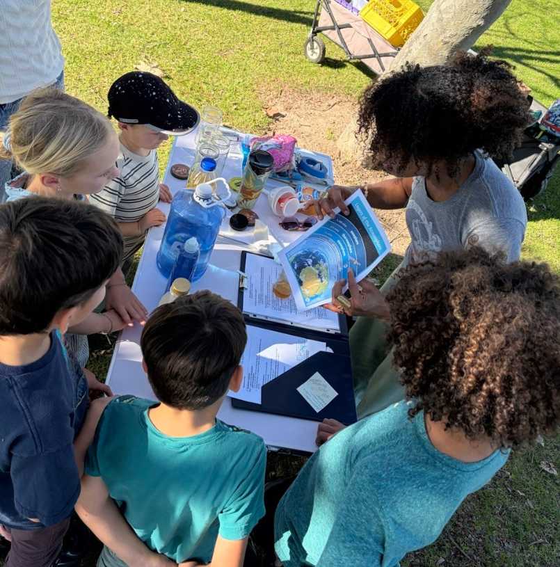 A group of children gathered around a table outdoors, listening to a woman holding a brochure. The table has books, papers, and water bottles. The scene takes place in a park with grass, a tree, and a cart in the background.