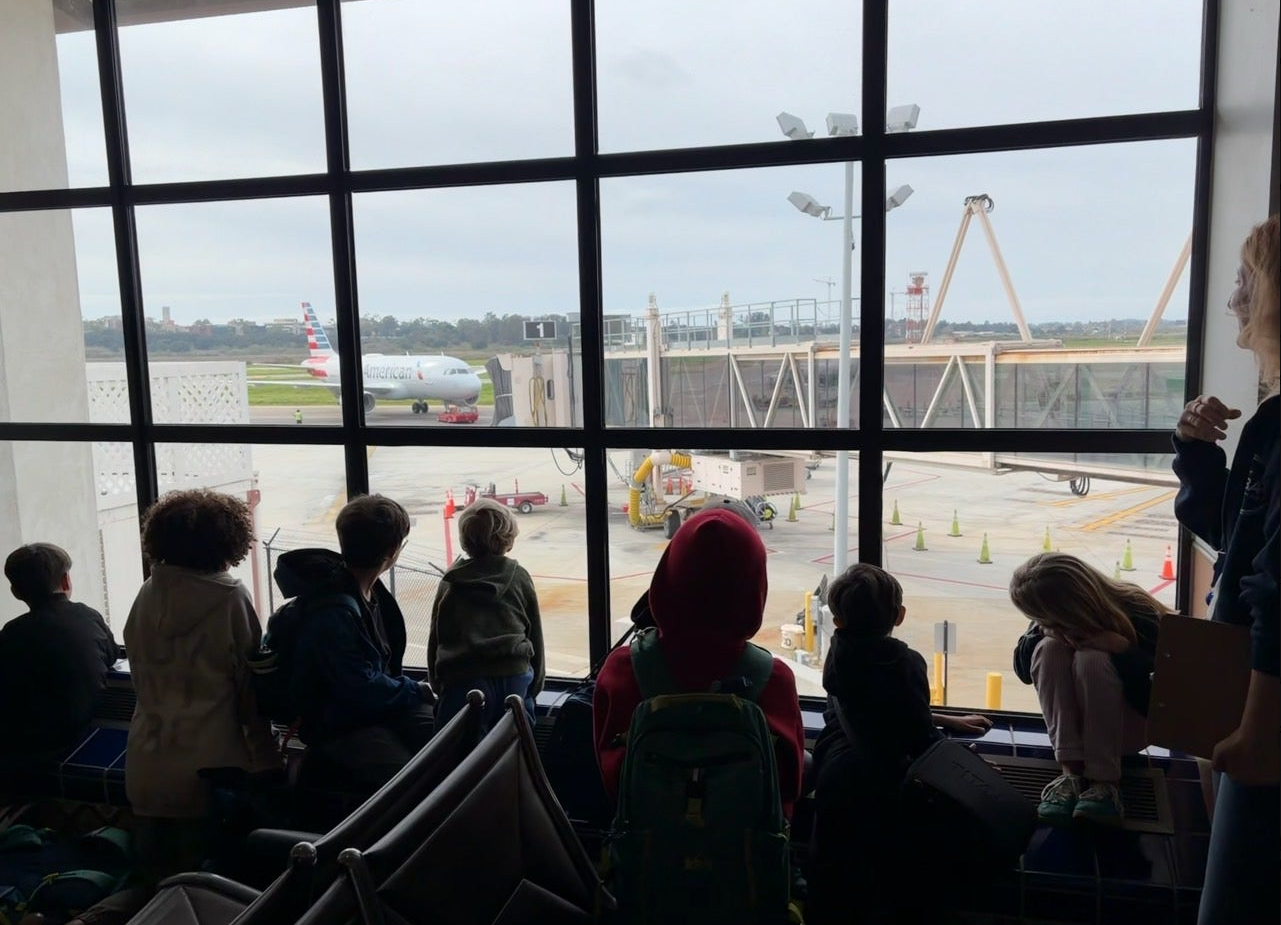 Children and adults sitting and standing by large airport window, watching American Airlines plane on tarmac outside.