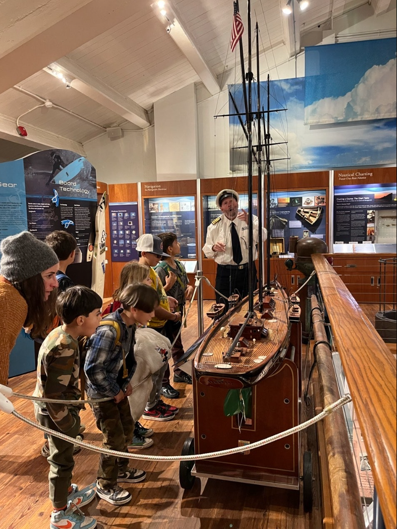 A group of children listening to a guide in a museum exhibit about ships. The guide, dressed as a ship captain, is talking about a model sailing ship on display.