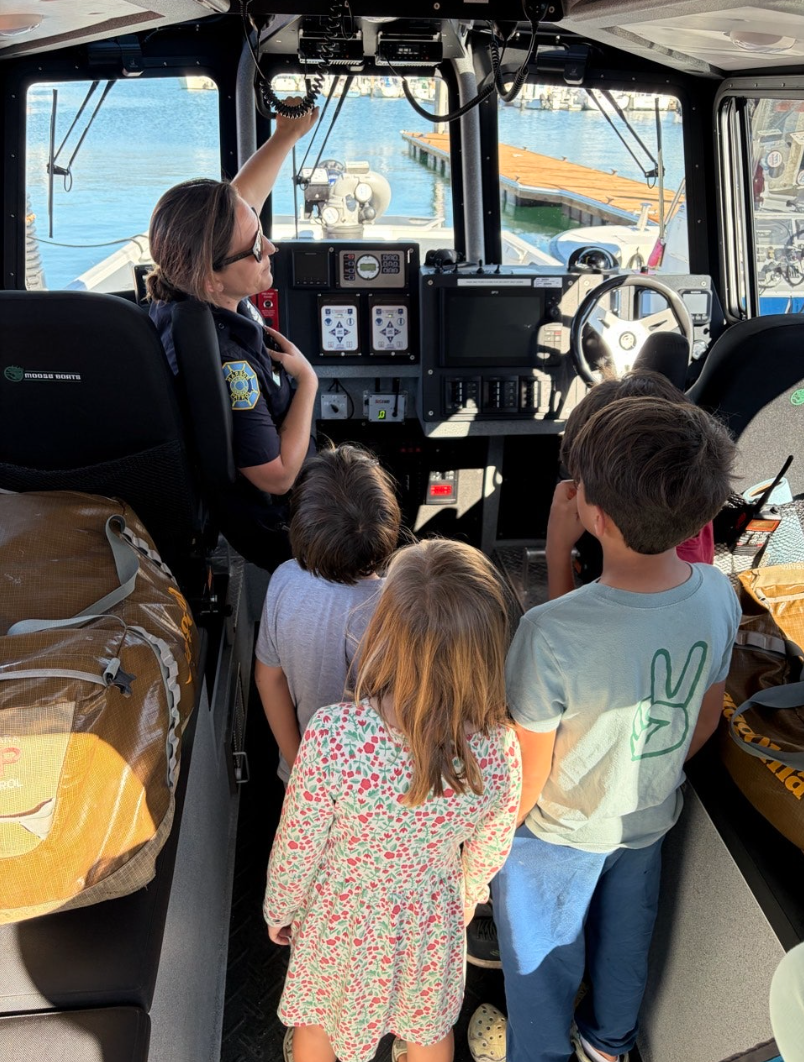 A police officer shows children the controls inside a boat's cockpit with water and a dock visible through the windshield.