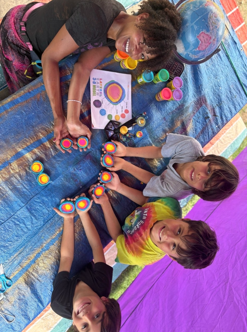 A woman and three children sitting at a table covered with paint and art supplies, making colorful tie-dye swirl patterns with cotton swabs. The woman is smiling and holding two swabs with rainbow designs, and a globe is visible in the background.