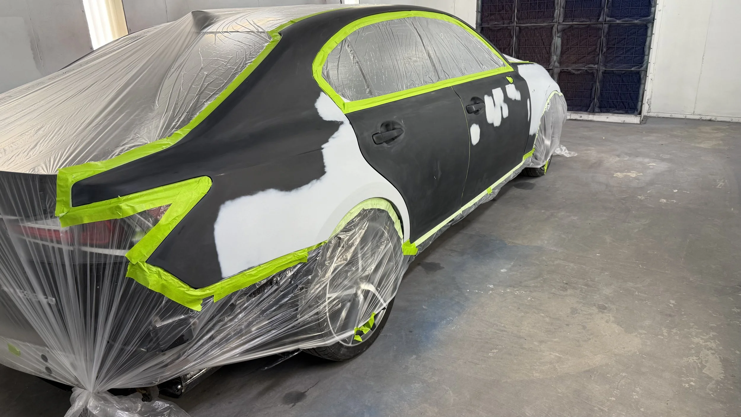 A car is covered with plastic sheeting and masking tape, with the body partially painted black and white, in a painting or repair shop.