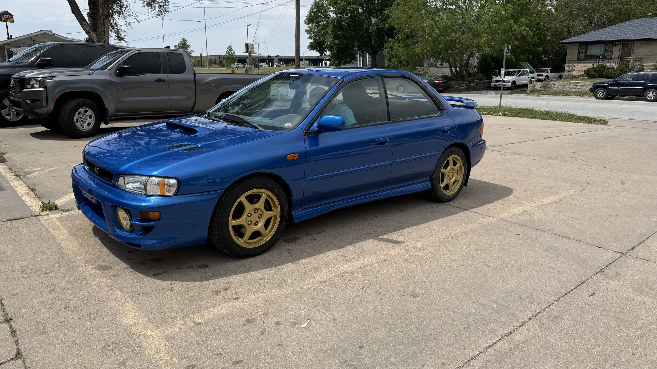 A blue sports car with gold wheels parked in a lot, with gray trucks and other vehicles in the background.