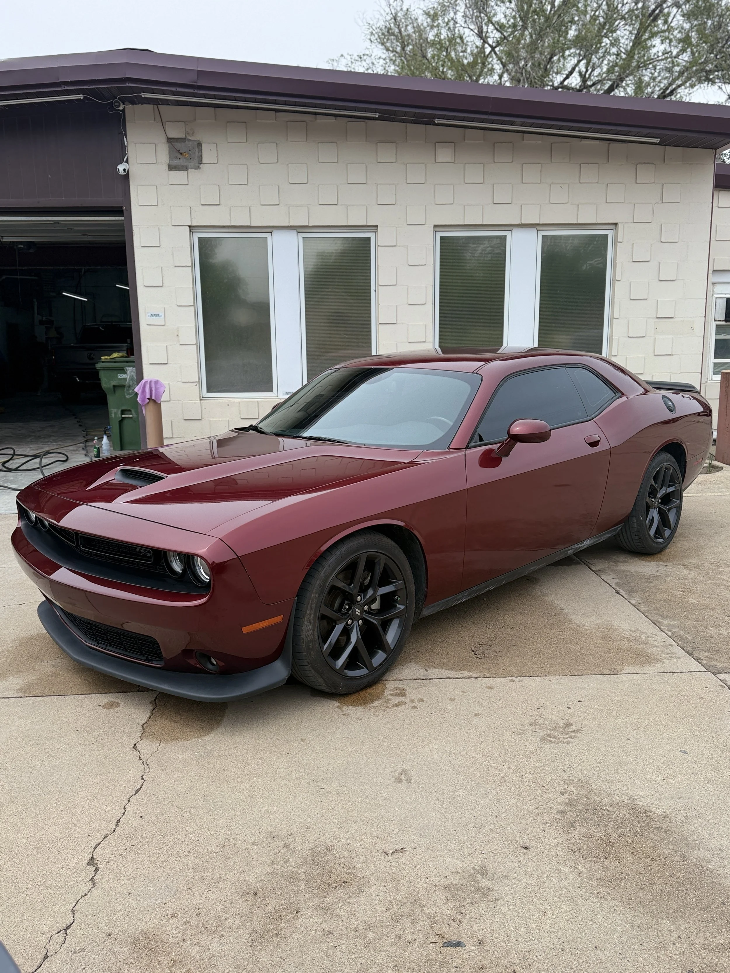 A red muscle car parked in front of a beige brick building with large glass windows. The building has a purple roof and a garage door on the left side. The ground is concrete with visible cracks.
