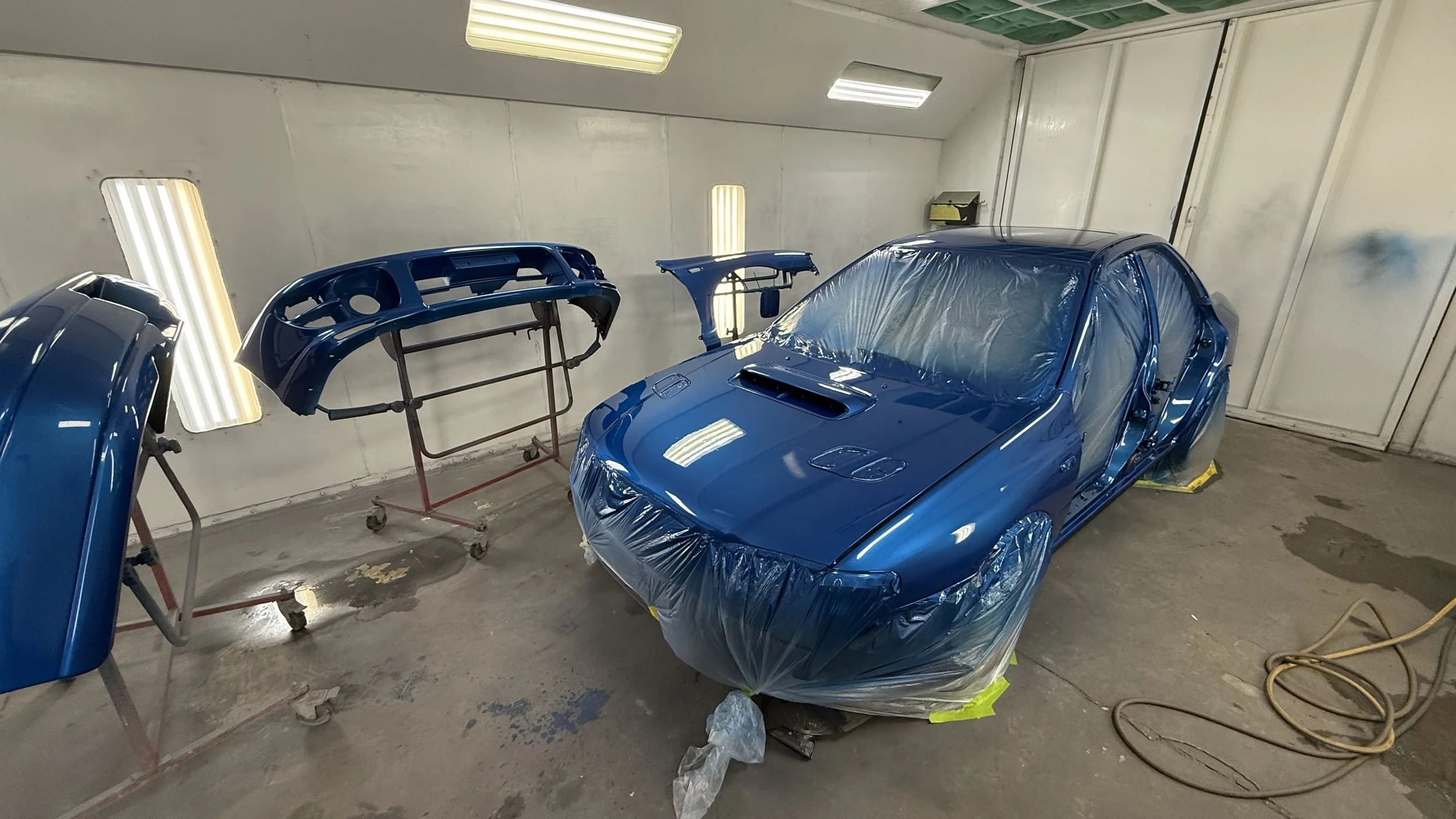 A blue car is being painted in a paint booth. The car's body is covered in plastic wrap, and the paint is fresh and shiny. Several car parts, including bumpers, are mounted on stands around the car for painting.