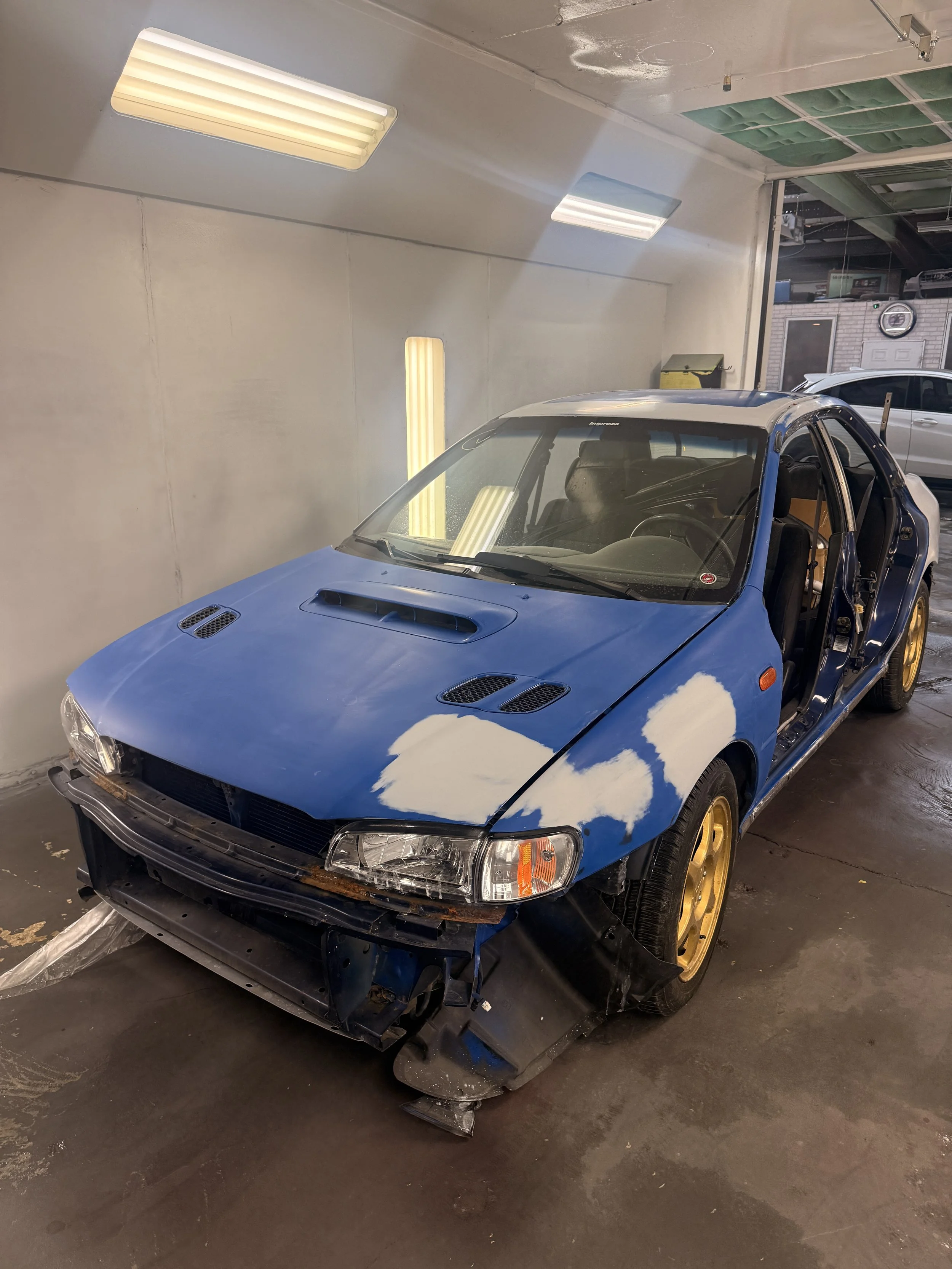 A blue car with yellow wheels is in a garage, with its front bumper and side panels removed, showing the inner structure. The car's roof has a patch of white paint and the windshield is still intact. The garage has white walls, fluorescent lights, and a partially open garage door revealing other vehicles and equipment.
