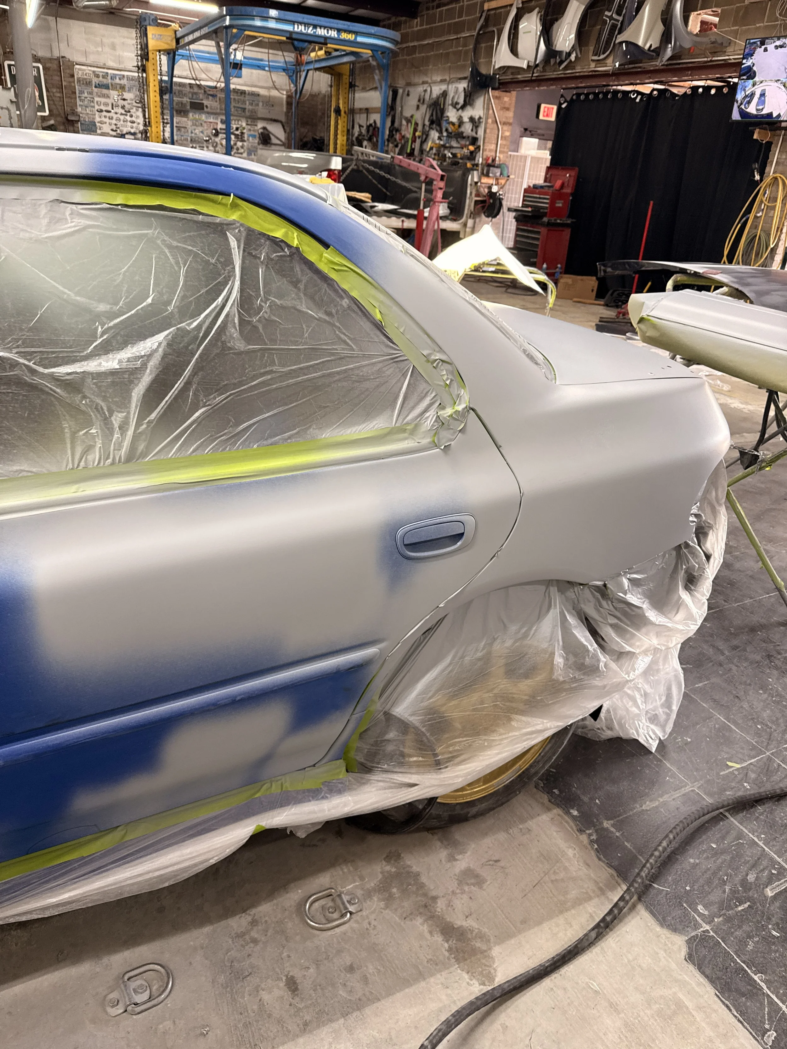 A silver car is being prepared for painting inside an auto body shop. The windows are covered with plastic sheeting, and yellow painter's tape is used to mask the edges. The surroundings include various tools, car parts, and equipment for auto repair.