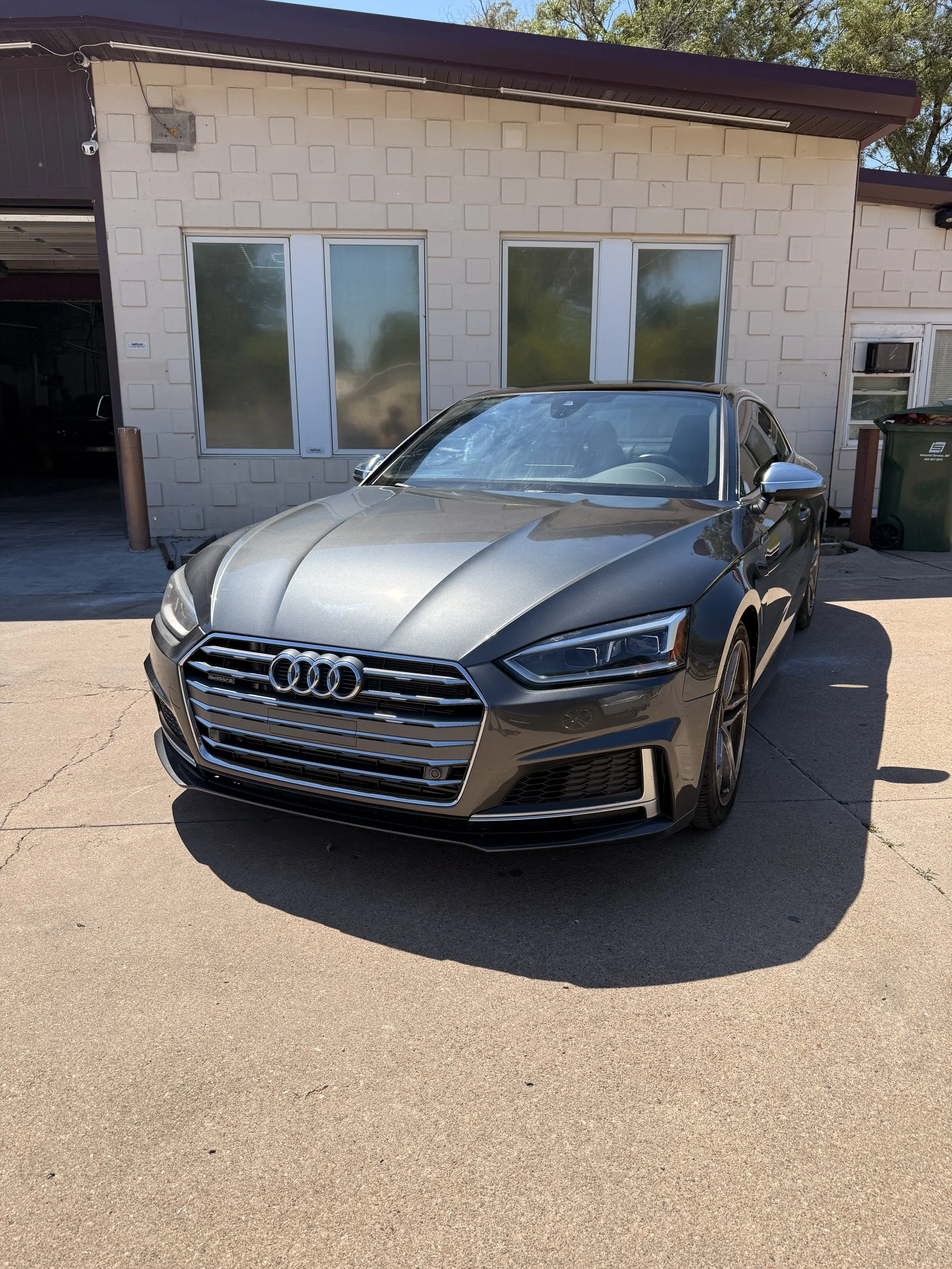 A silver Audi sedan parked in front of a beige brick building with three large windows and one smaller window. There is a garage door to the left and a green trash bin to the right. The sky is clear and blue.