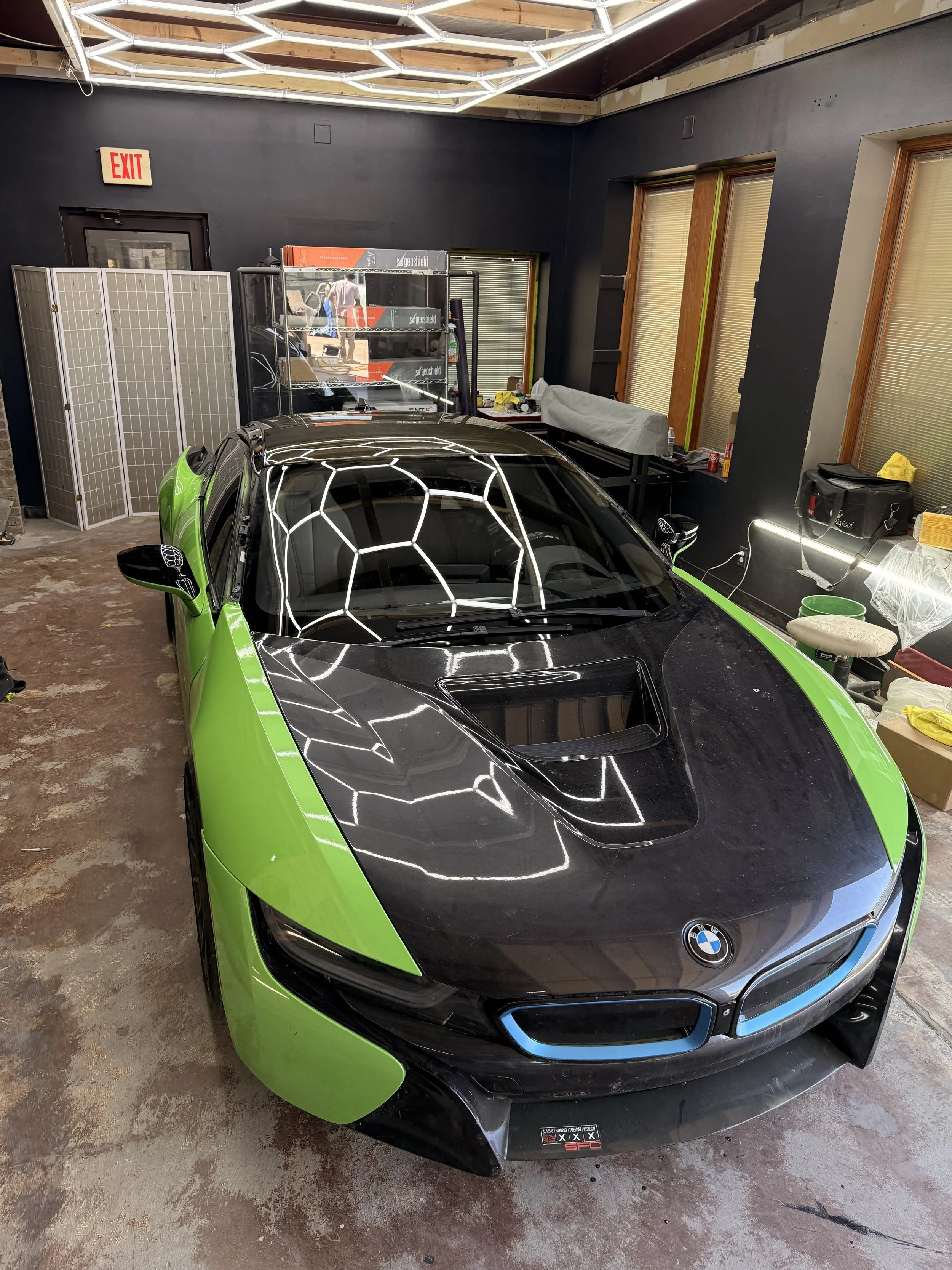 A black and green BMW sports car parked inside a garage, with reflective ceiling lights, an exit door, window blinds, and various tools and equipment on the shelves and tables.