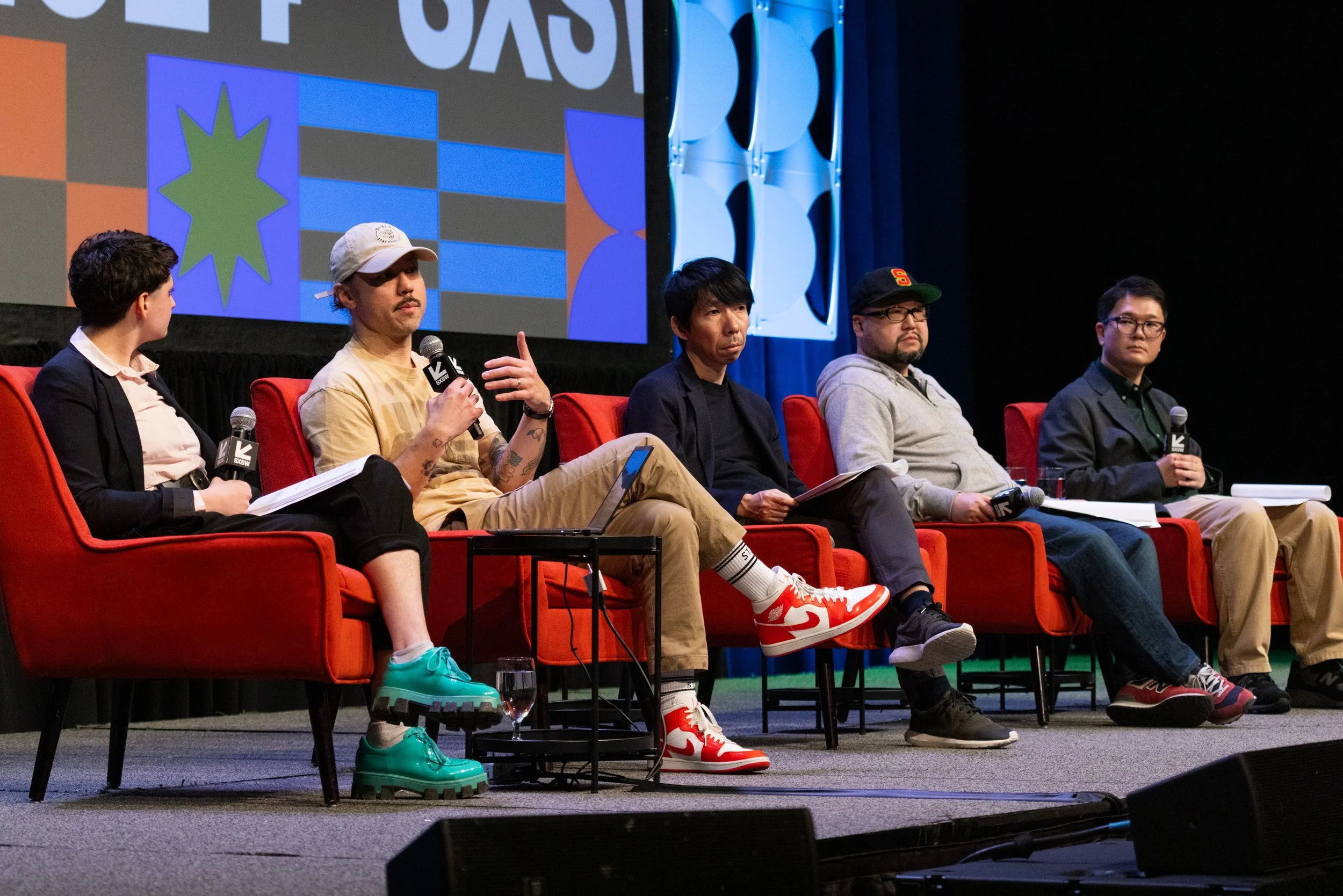 Panel of five diverse men sitting on a stage with a large screen behind them, participating in a discussion at an event. Each man holds a microphone, and there are blank papers or notebooks on their laps. The men are dressed casually, and the stage has red chairs, with a dark backdrop and abstract blue and white designs on the panel wall.