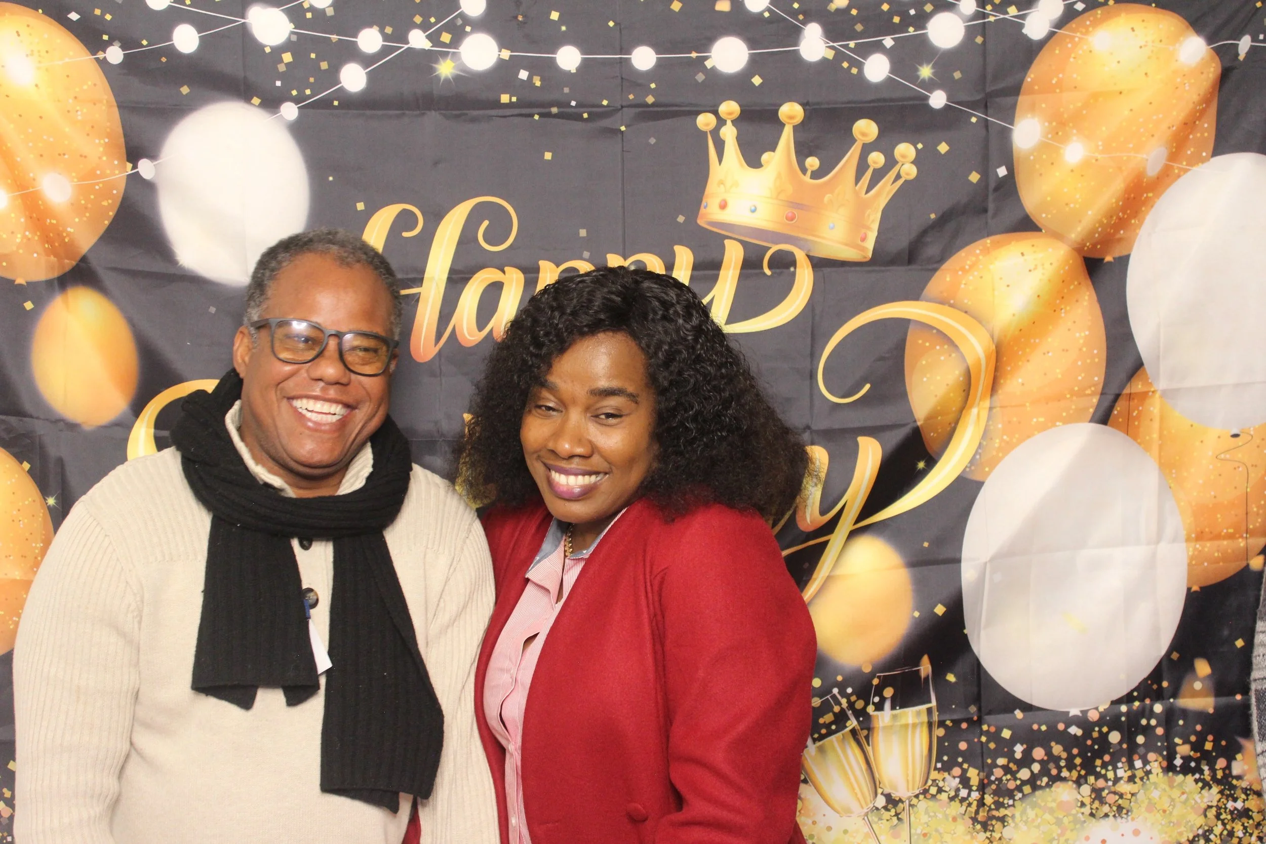 Two people, a man and a woman, smiling at a celebration event with a black and gold themed backdrop, balloons, confetti, a crown graphic, and text that reads "Happy Birthday".