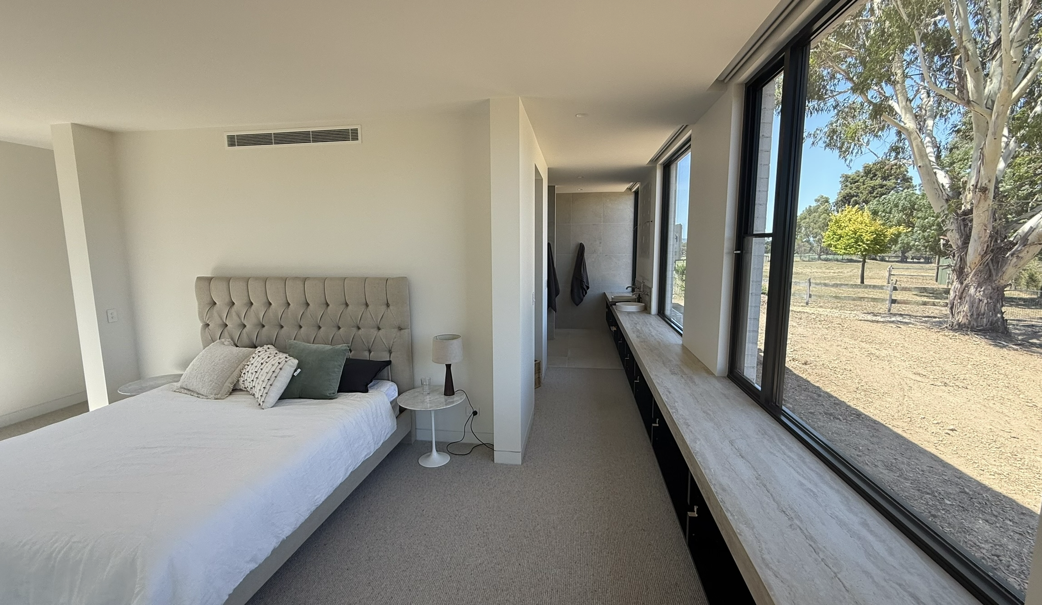Modern bedroom with large window overlooking outdoor landscape, bed with tufted headboard, side table with lamp, and built-in countertop along window.