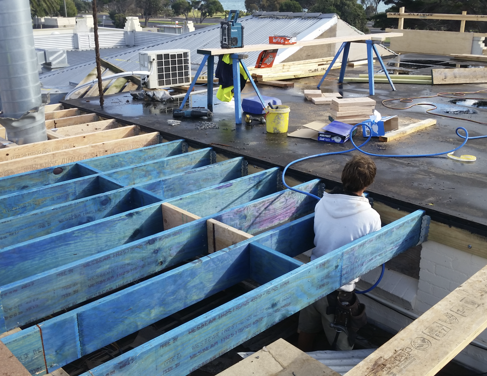 A construction worker sitting on the edge of a building under construction, with a blue wooden frame in front of them. The roof is partially finished with various construction tools, materials, and equipment scattered around.