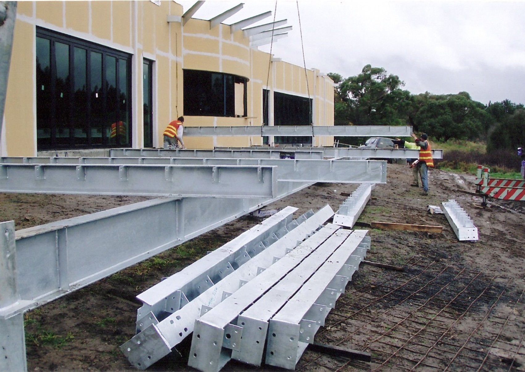 Construction workers assembling steel framework for a building, with some workers wearing safety vests, on an overcast day, with trees in the background.