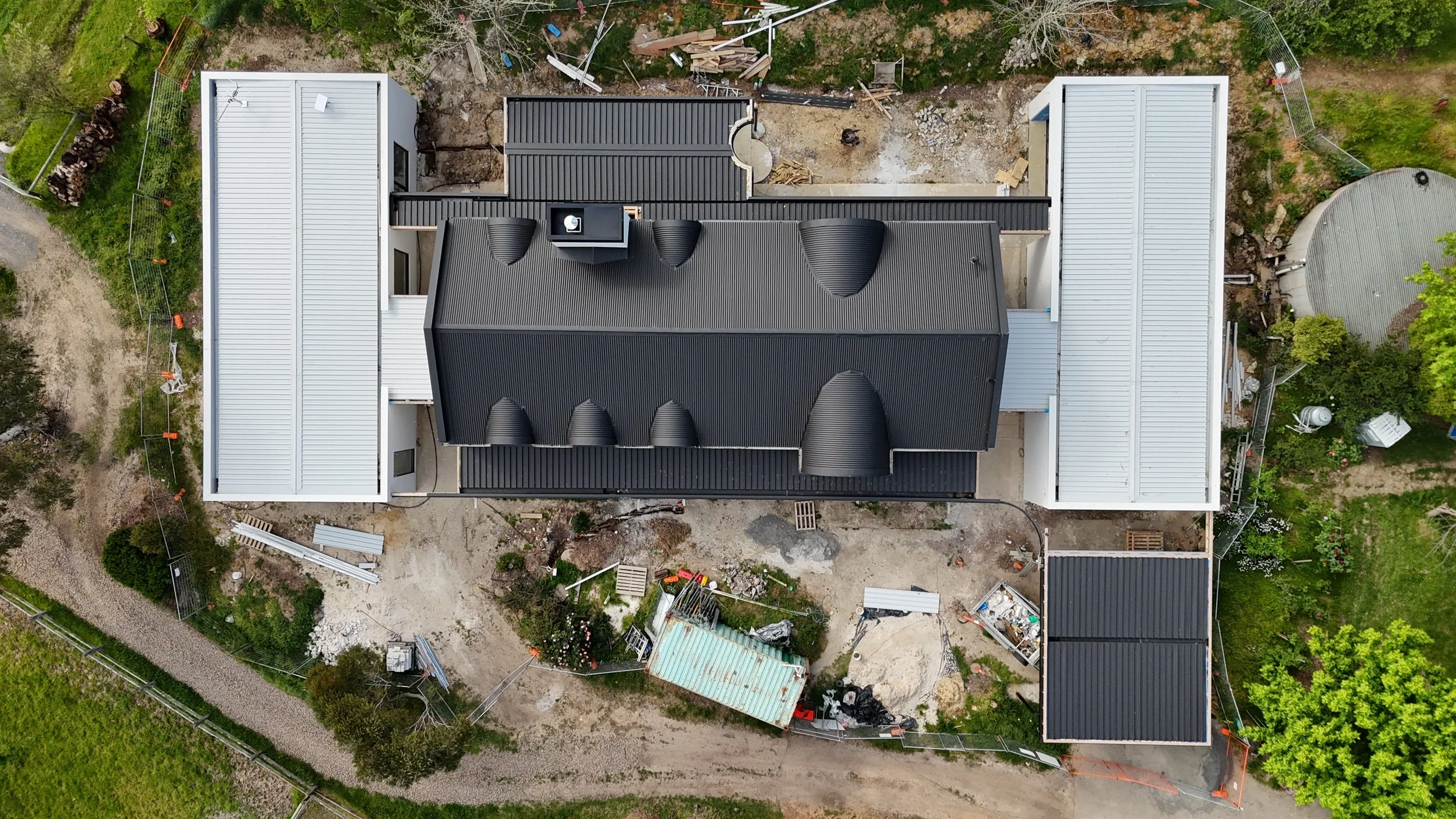 Construction site of a modern building with a white exterior, large arched windows, and wooden accents. The area around the building is unfinished with construction materials, dirt, and gravel.