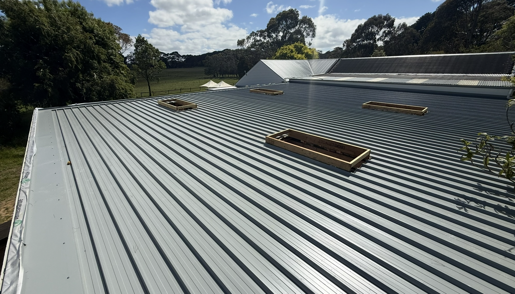 Metal roof with multiple rectangular skylight frames installed, surrounded by trees and a partly cloudy sky.