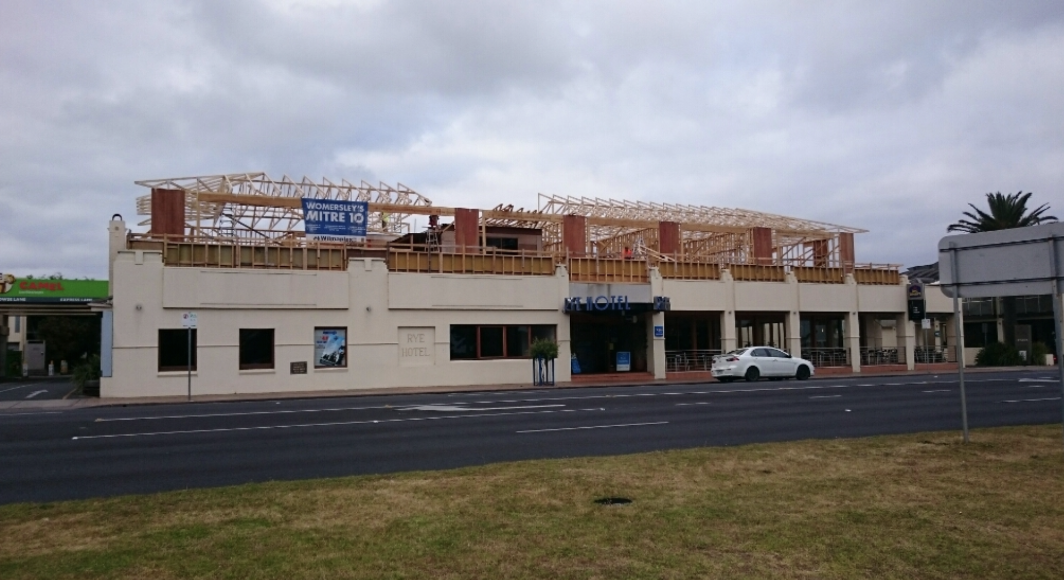 The Rye Hotel under construction with wooden framing on the upper floors, parked car in front, cloudy sky, and a patch of grass and pavement in the foreground.