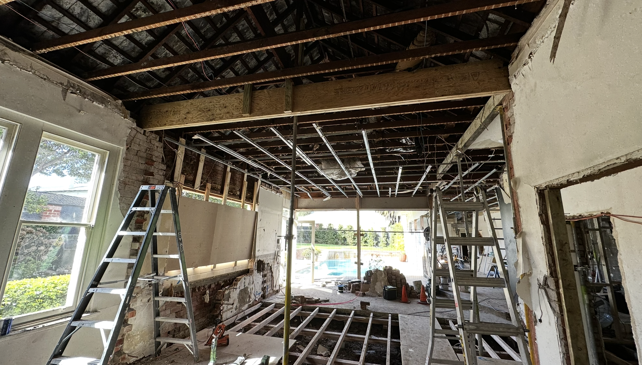 Interior view of a house under renovation, showing exposed ceiling beams, drywall removal, ladders, construction tools, and a partially open wall, with a backyard pool visible through the window.