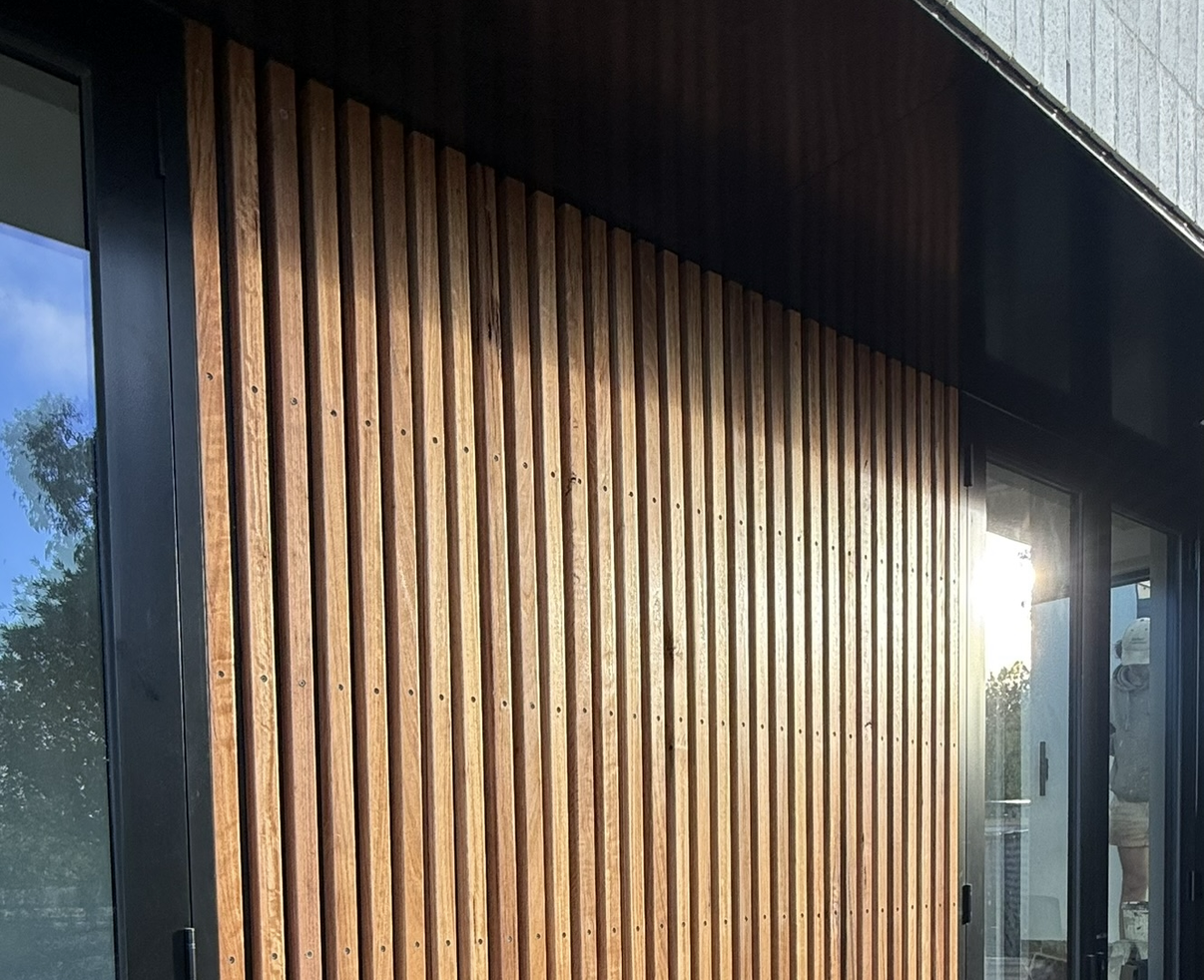 Exterior view of a building with vertical wooden slats and glass doors, reflecting the sky and trees.