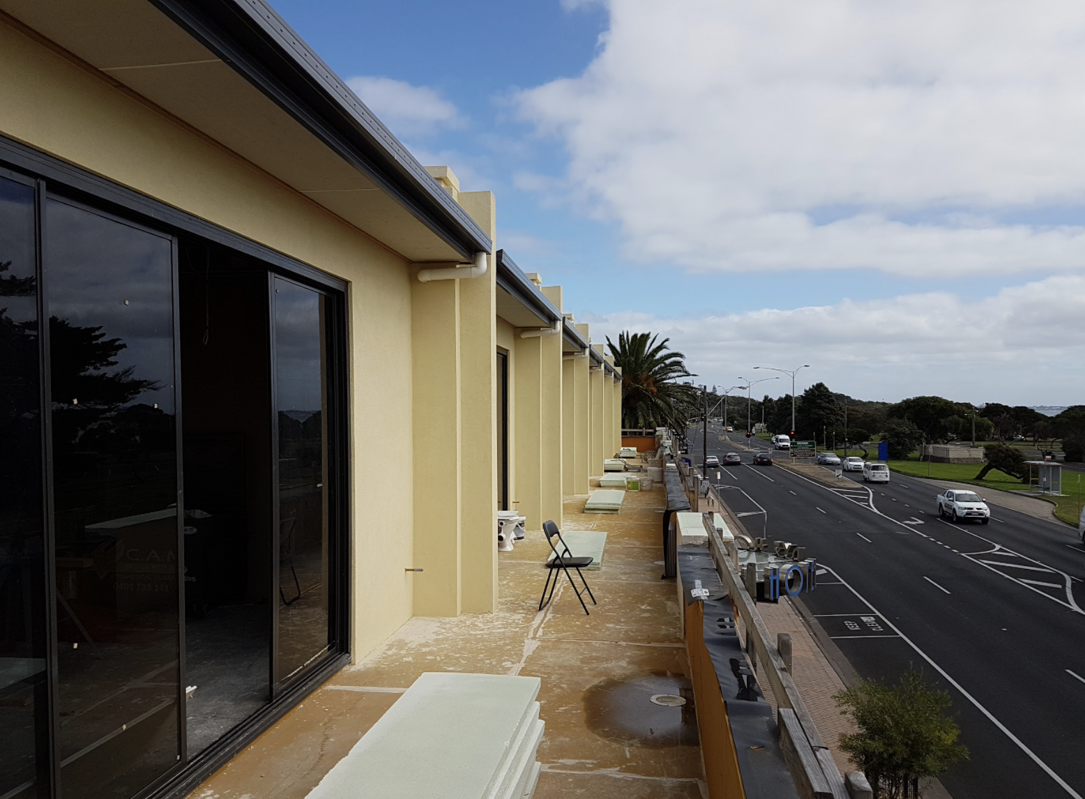 View of a balcony with yellow walls and sliding glass doors overlooking a busy street with cars and palm trees.