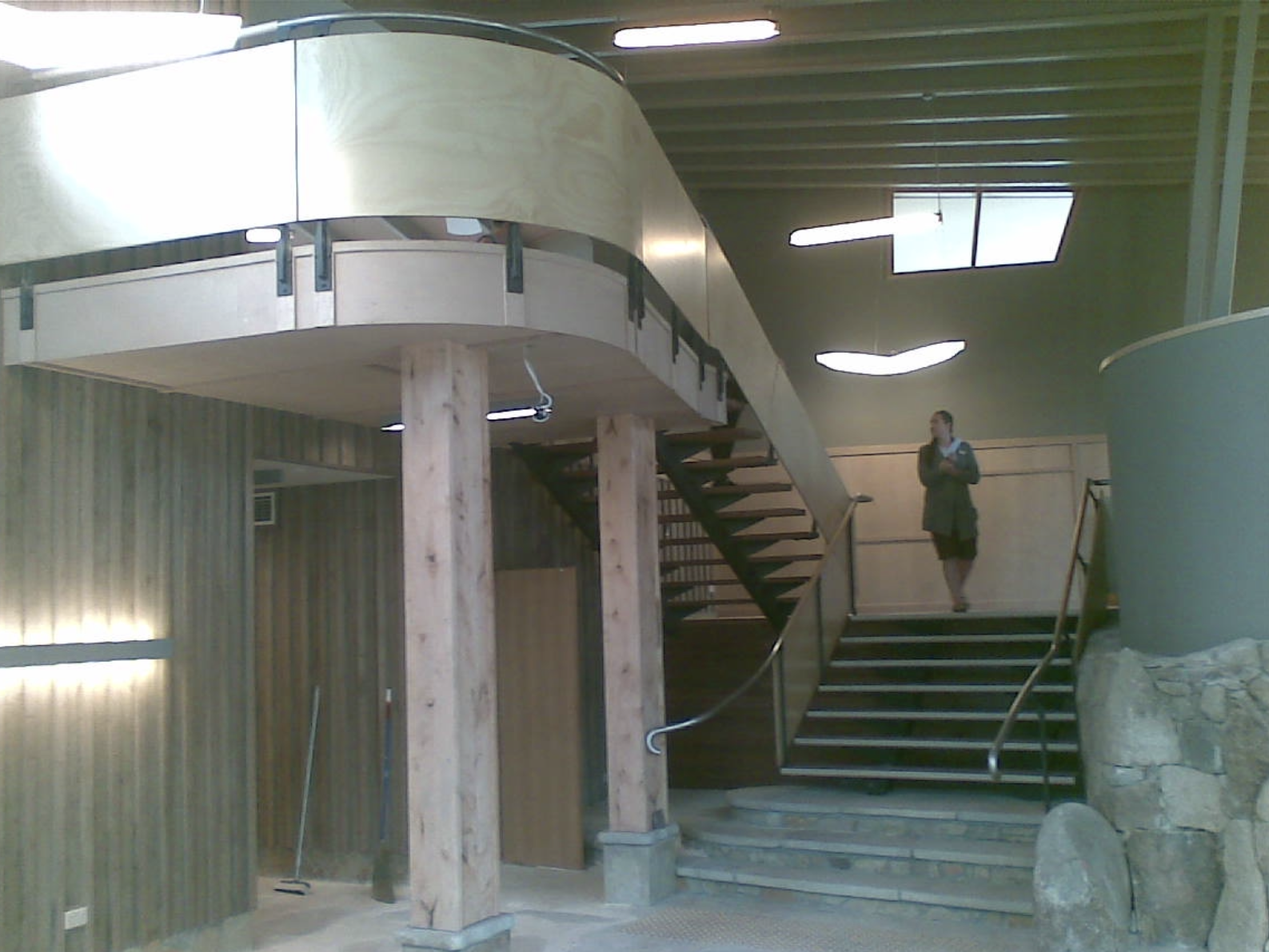 Interior of a modern building with a curved staircase, wooden pillars, and a woman standing on the stairs.