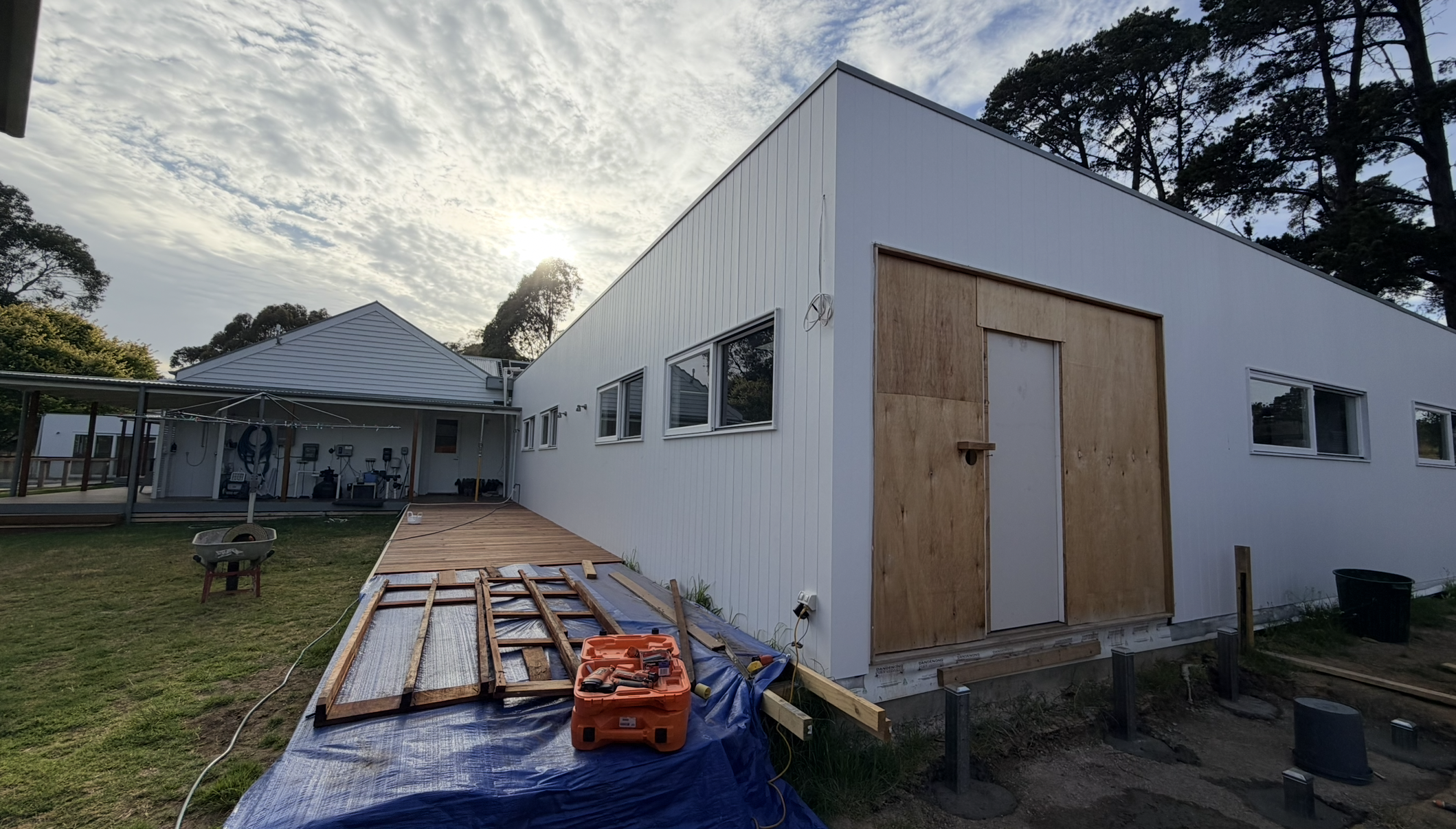 Under construction modern white house with wooden door frame, construction tools, and materials on the ground. The sky is partly cloudy with the sun setting behind trees.