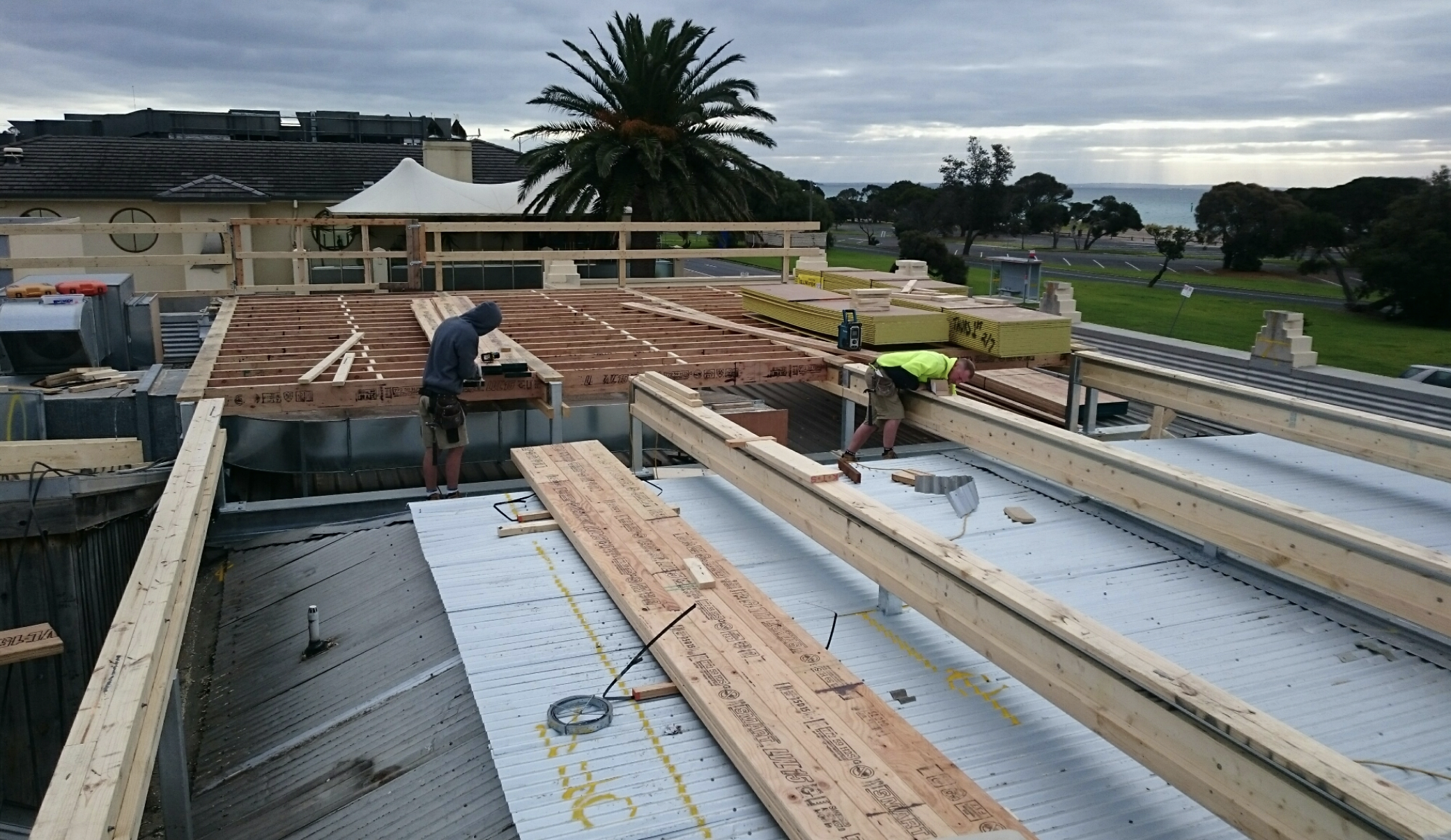 Construction workers building a rooftop with wooden framing and metal roofing, overlooking a park and water body.