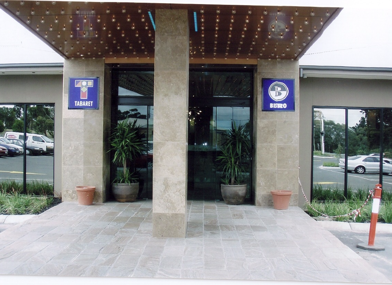 The entrance to a building with two blue signs reading 'Tabaret' and 'Bistro'. Potted plants are placed on either side of the glass doors, and there is a sidewalk and parking lot with cars outside.