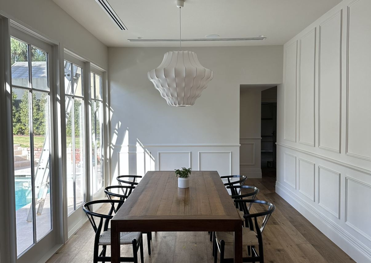 A modern dining room with a wooden table, six black chairs, a white decorative light fixture, white paneled walls, and large glass doors leading to an outdoor area with a pool.