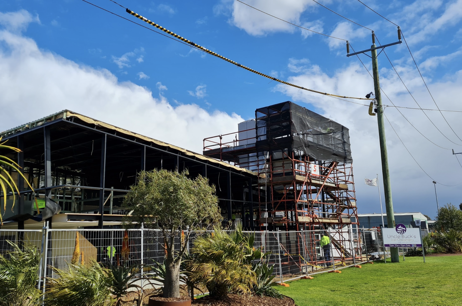Construction site of a building with scaffolding, some workers, and trees in front, under a partly cloudy blue sky.