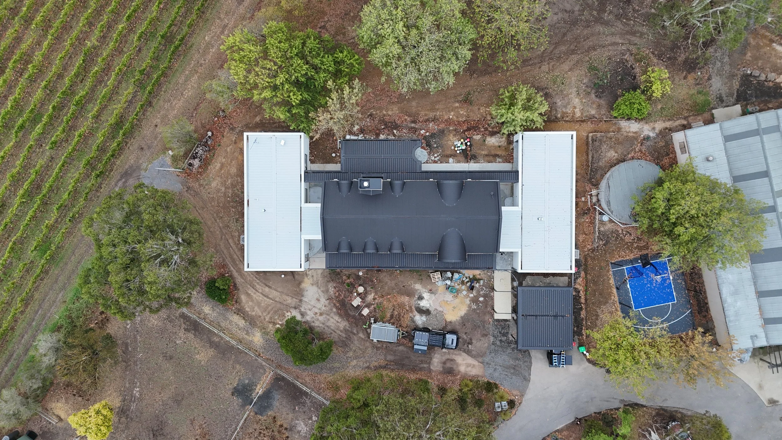 Aerial view of a house with a black roof, surrounded by trees, a trampoline, and a shed, with a dirt driveway and patches of grass.