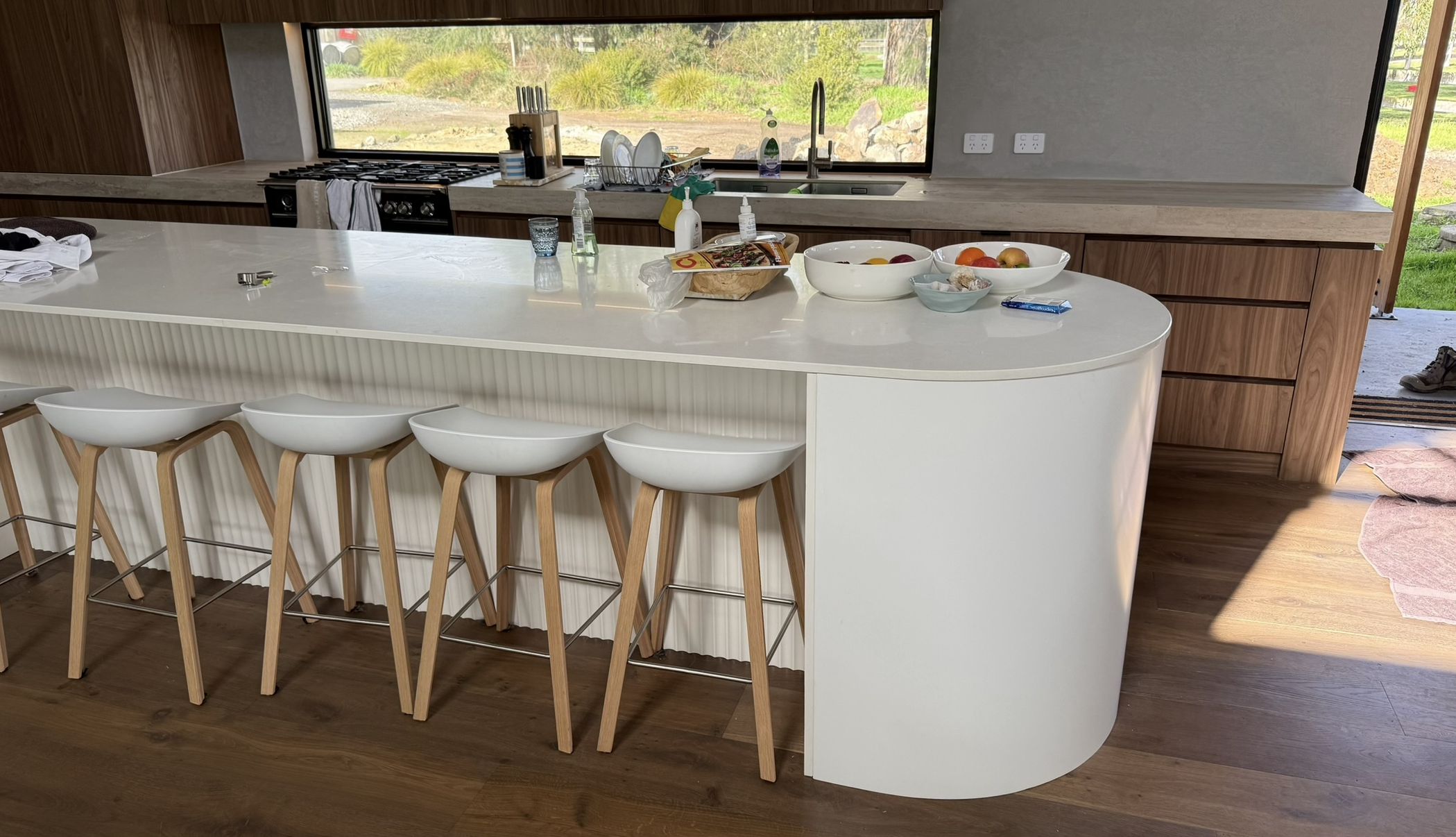 Modern kitchen with a large white island countertop, four white bar stools with wooden legs, a sink with a window behind it, and various kitchen items and bowls on the counter.