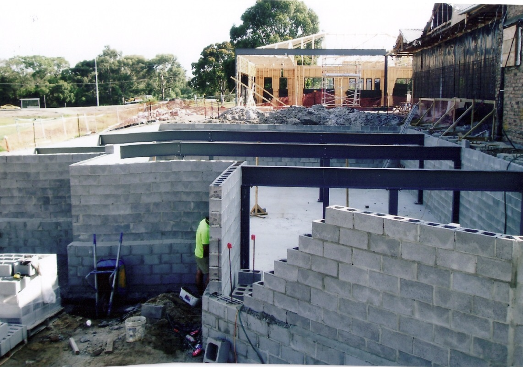 Construction site with workers building brick walls for a new building, with steel beams in place and a partially constructed wooden roof structure in the background.