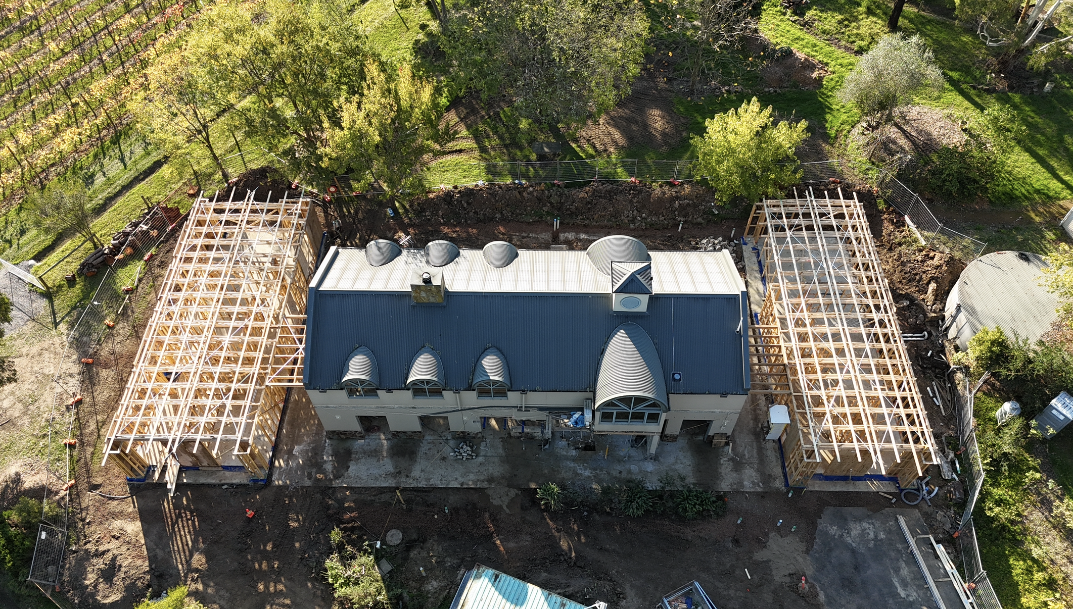 Aerial view of a building under construction with wooden framing on both sides, a metal roof, and surrounded by green trees and a fenced yard.